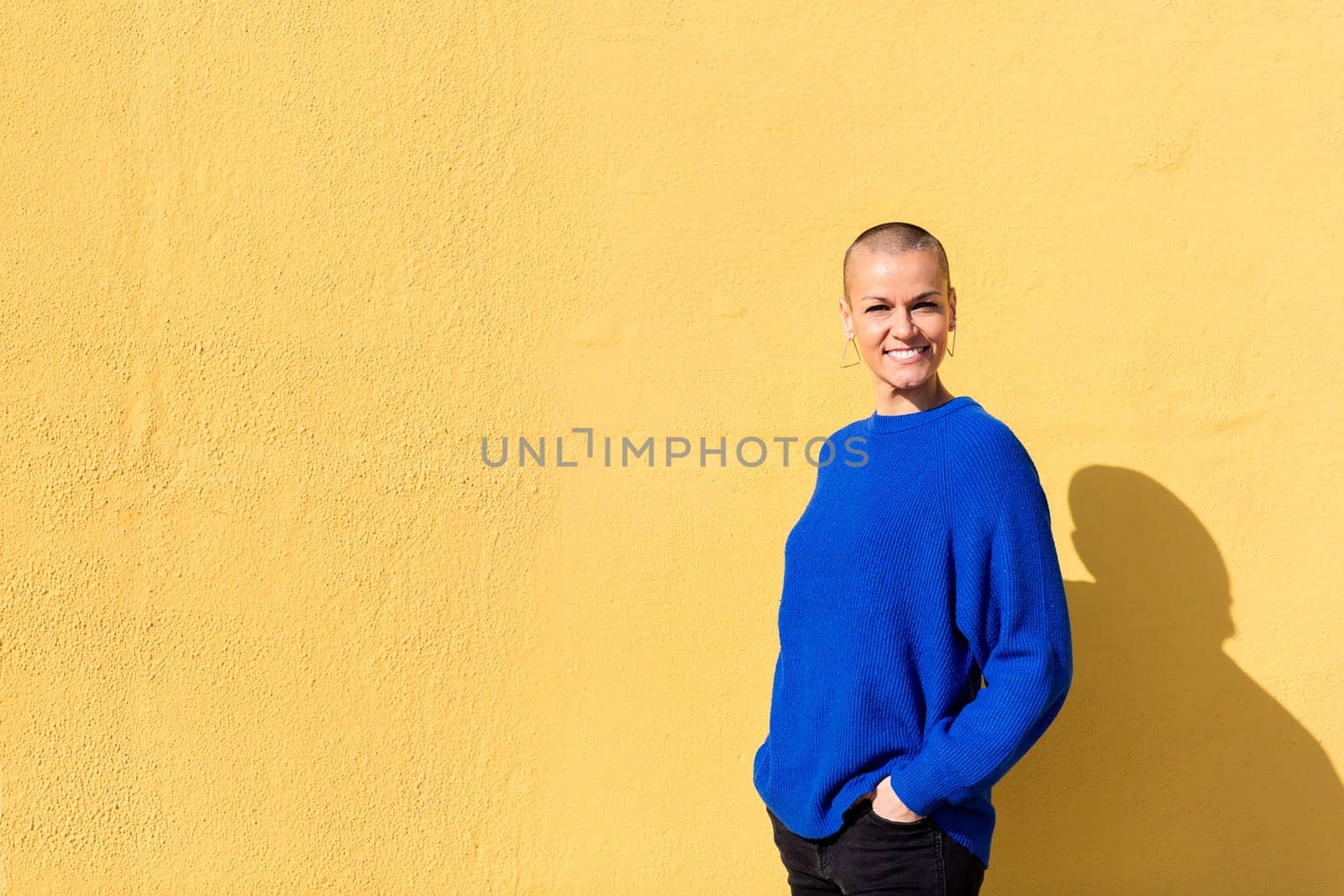 woman with a shaved head and a blue sweater smiling looking at camera with a yellow wall in the background, copy space for text