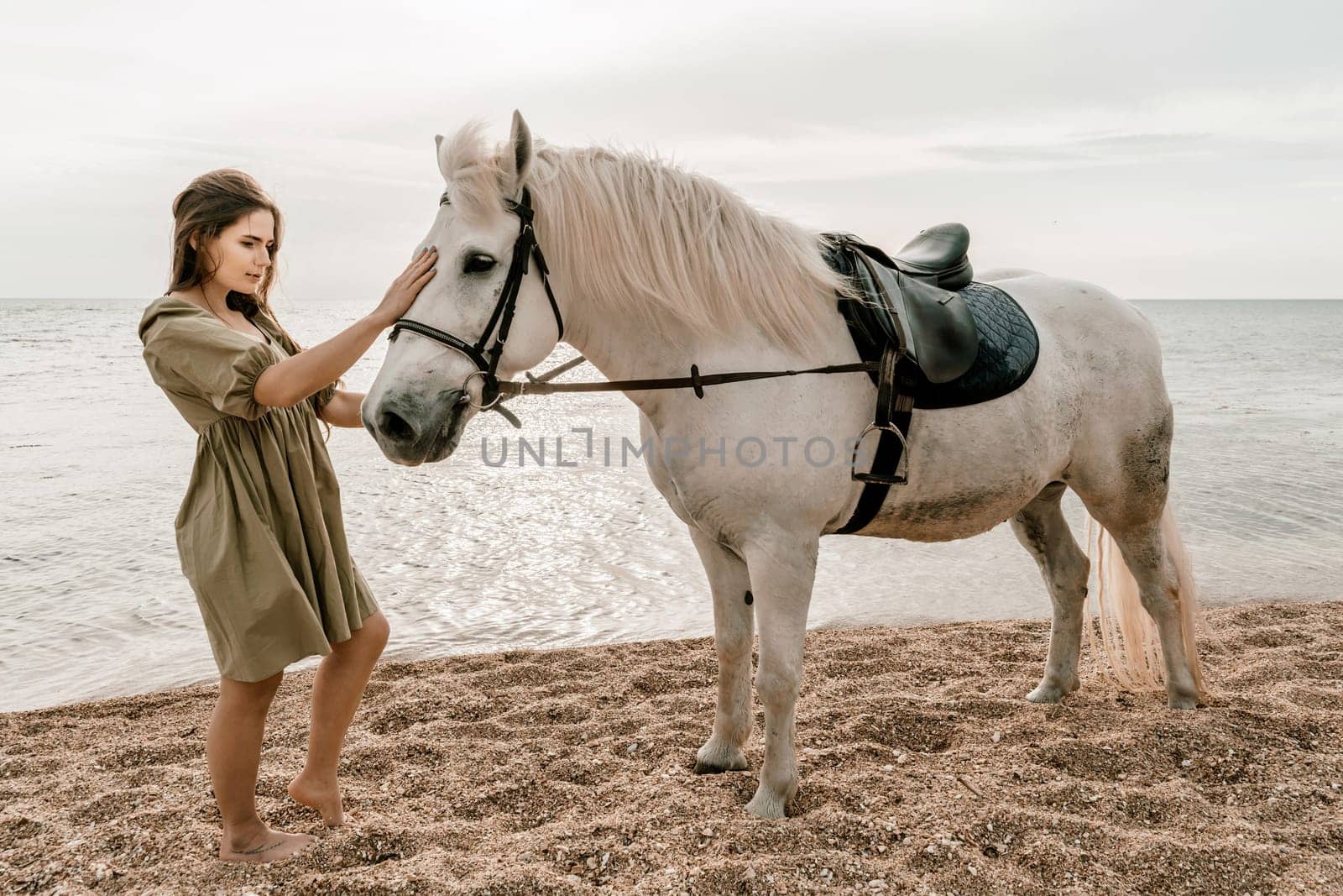 A woman in a dress stands next to a white horse on a beach, with the blue sky and sea in the background