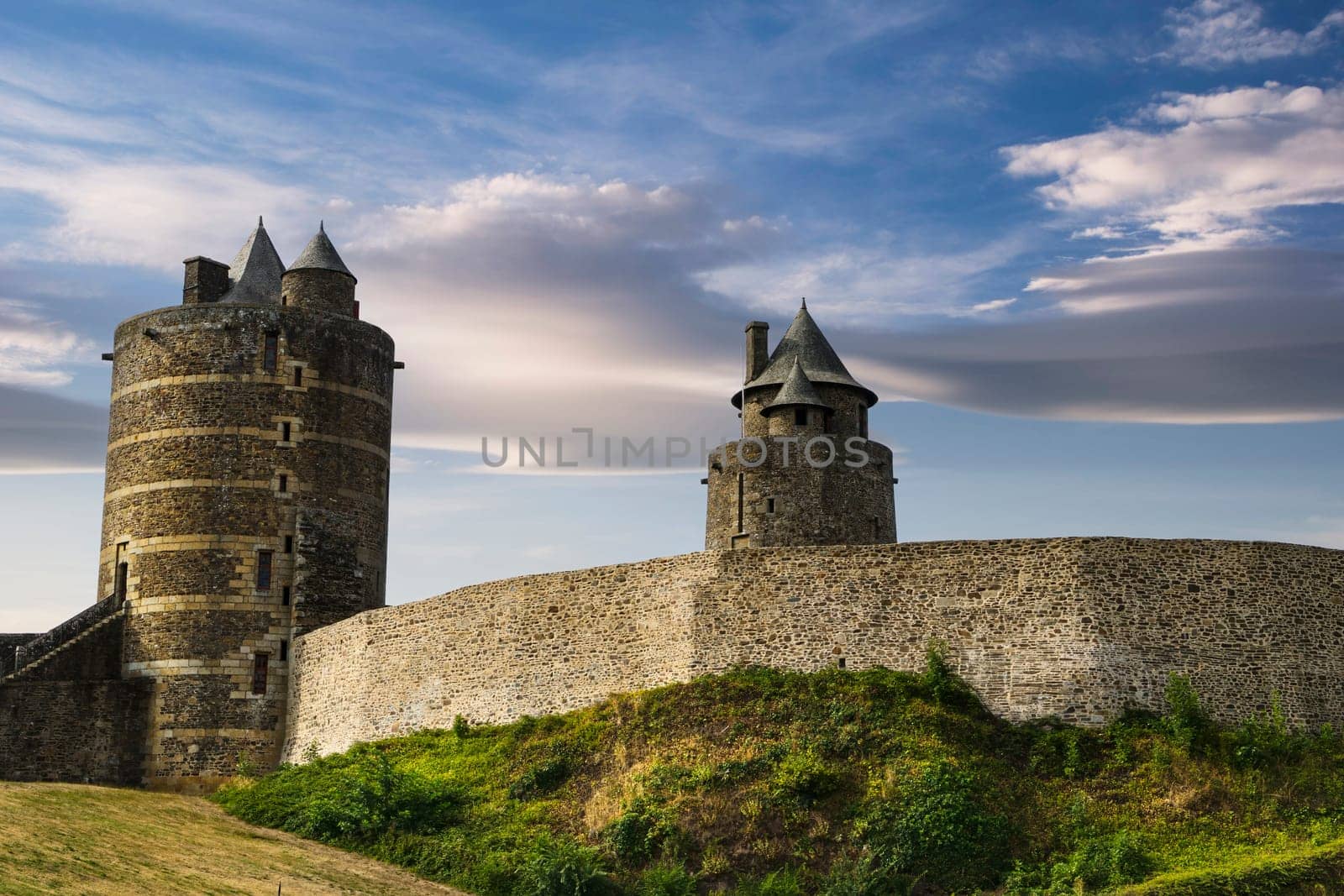 View of the wall and towers of the medieval castle of the French town of Fougeres built in the 13th century, with a background of blue sky and clouds at sunset.