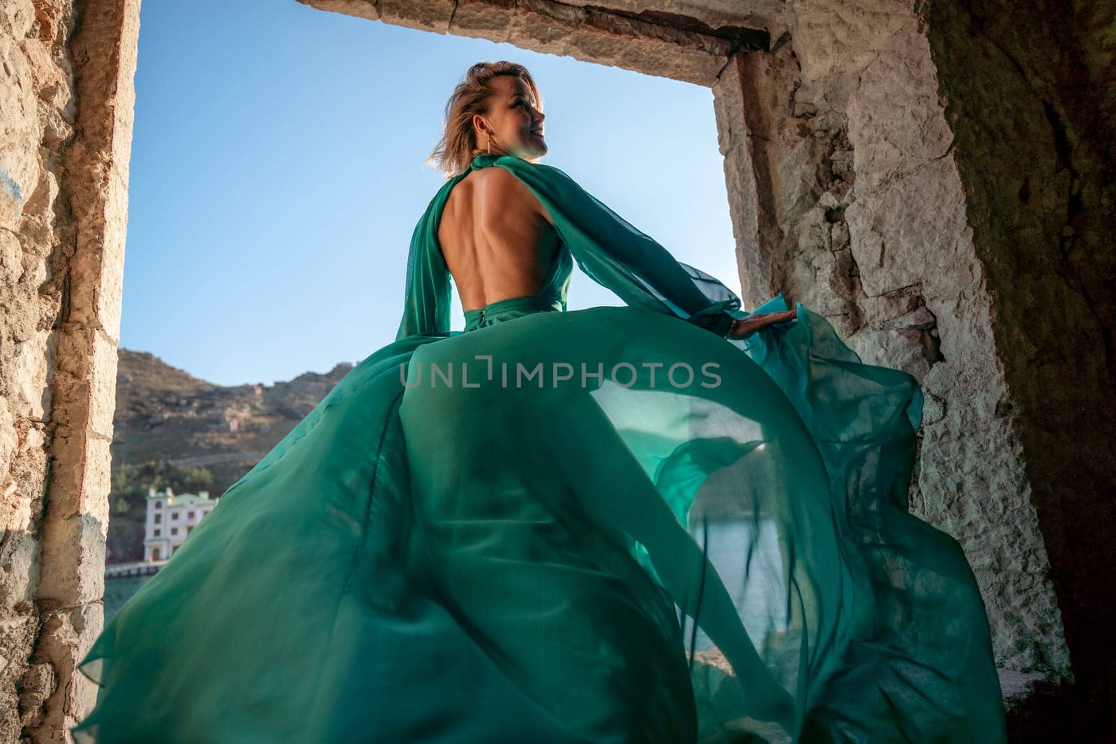 Rear view of a happy blonde woman in a long mint dress posing against the backdrop of the sea in an old building with columns. Girl in nature against the blue sky