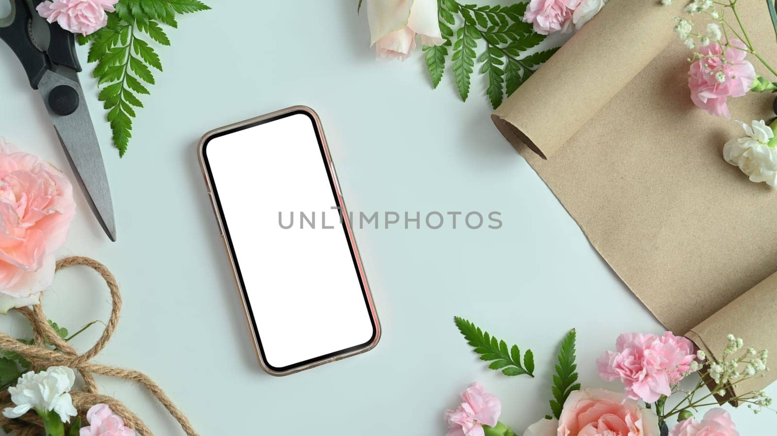 Smartphone with blank screen, pink flowers, shears, wrapping paper and leaves arrangement on white background.