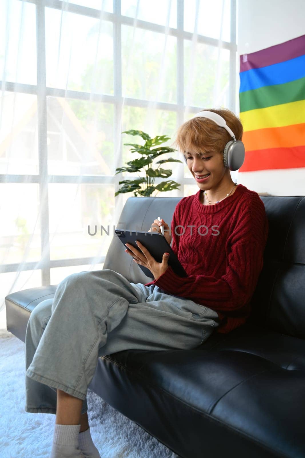 Attractive young gay man wearing headphone using digital tablet, surfing social media, watching movies while relaxing on couch at home.