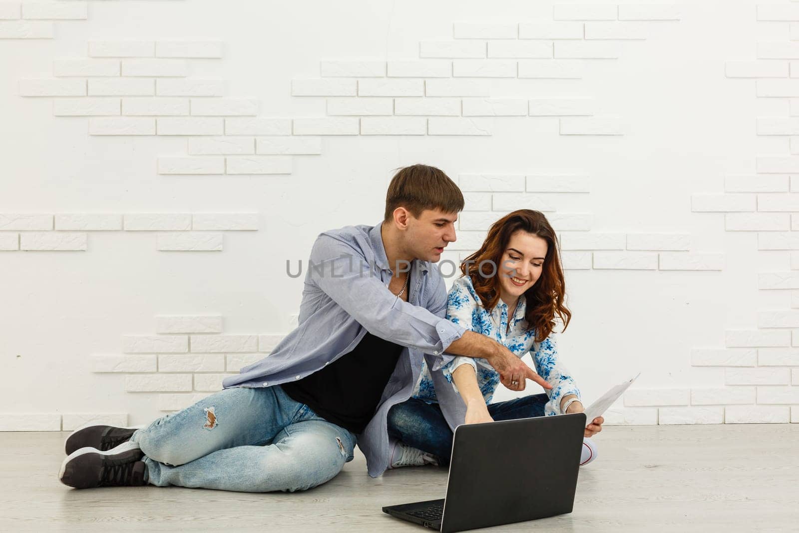 Couple Use Laptop Computer, while Sitting on the Living Floor room of their Apartment. Boyfriend and Girlfriend Talk, Shop on Internet, Choose Product to Order Online, Watch Streaming Service. by Andelov13