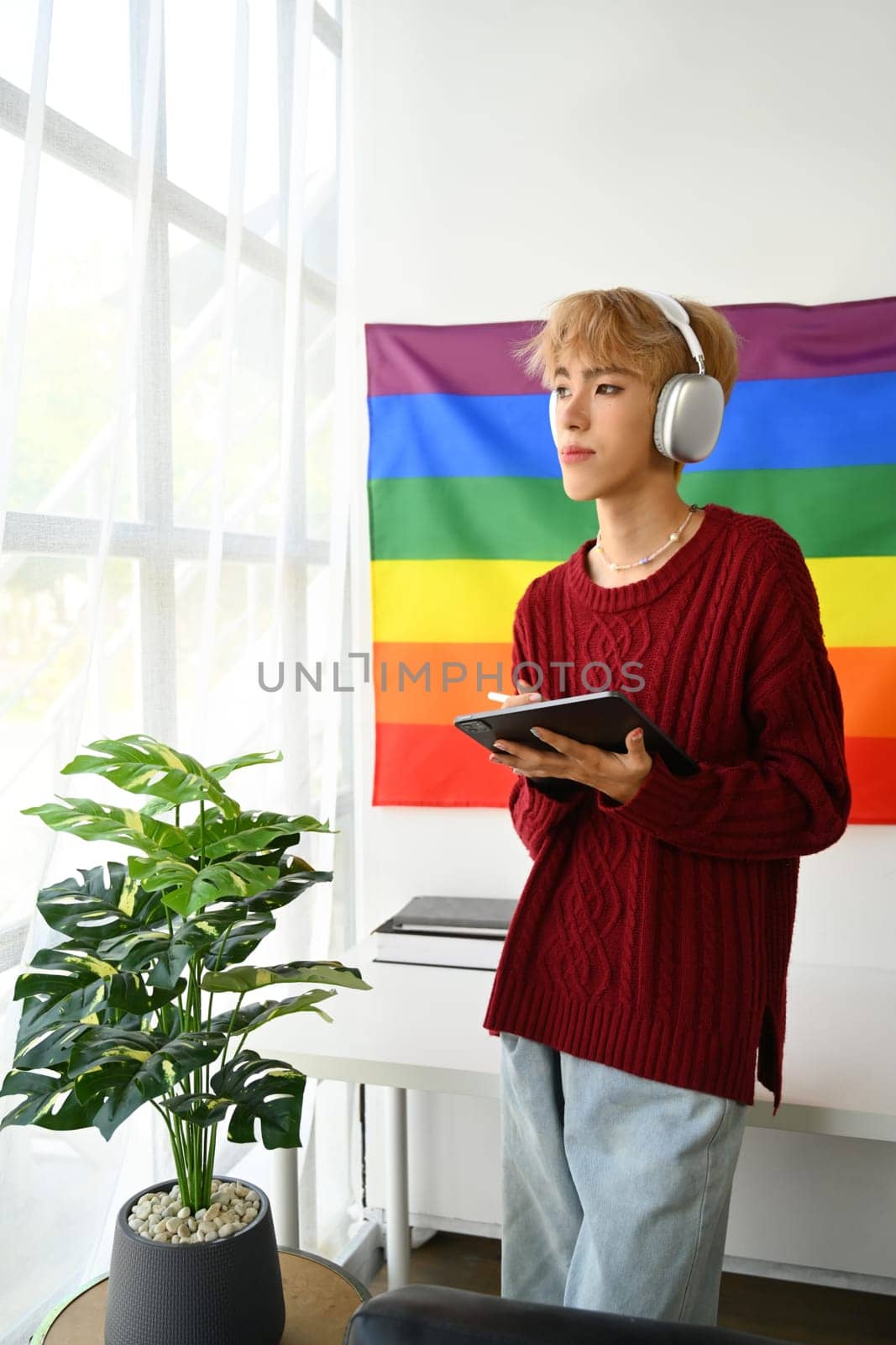 Portrait of thoughtful young Asian gay man standing in cozy living room with pride rainbow flag and looking out of window.