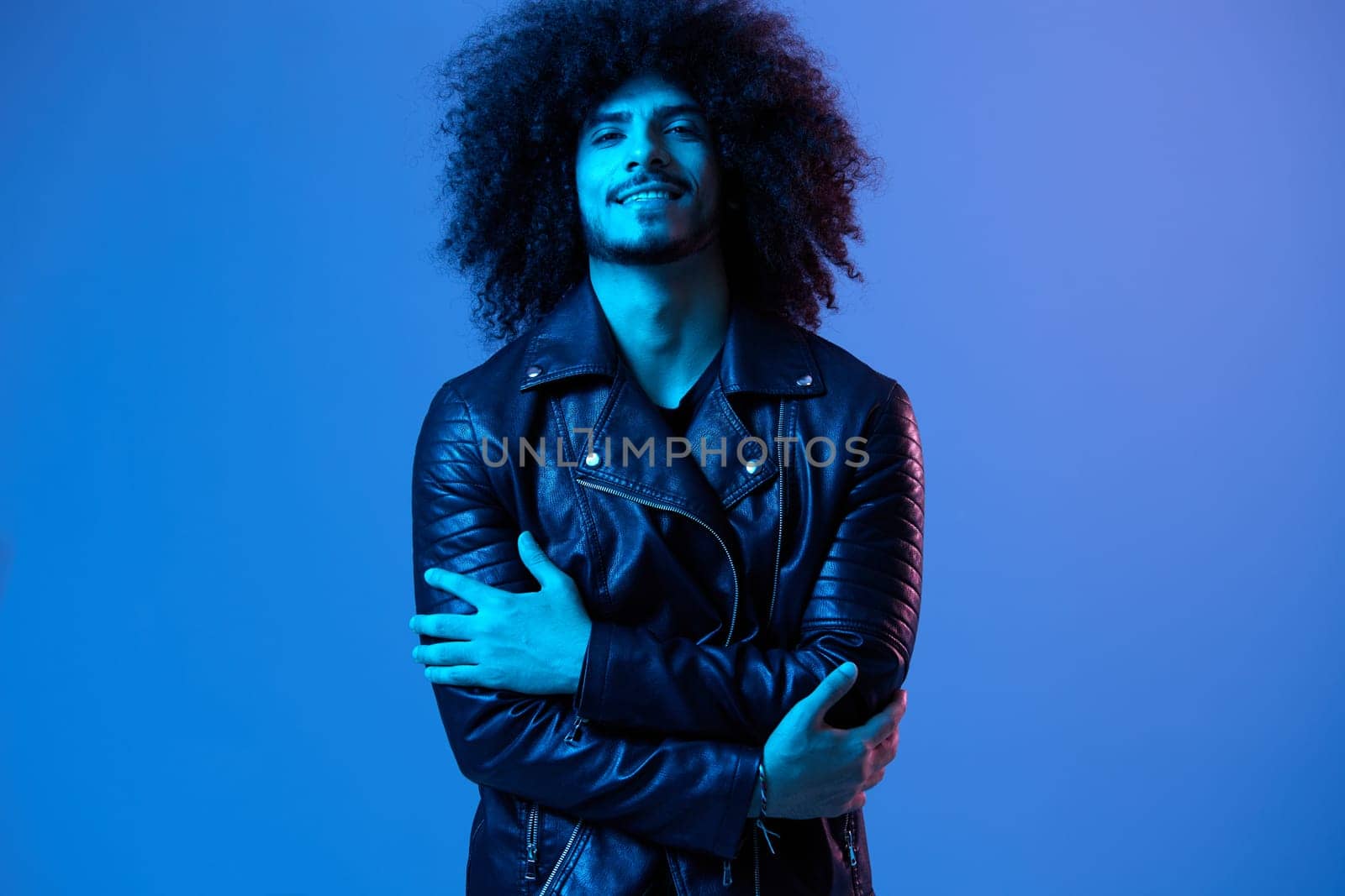 Portrait of fashion man with curly hair on blue background multinational, colored light, black leather jacket trend, modern concept. High quality photo