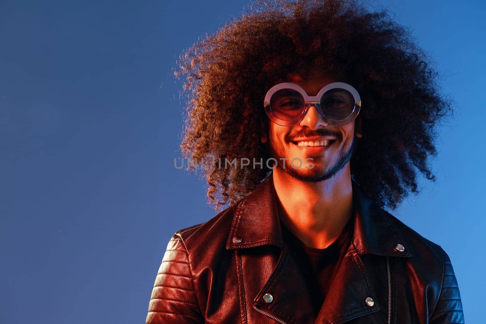 Portrait of a stylish man with curly hair with glasses smile on blue background multinational, color light, black leather jacket trend, modern concept. by SHOTPRIME