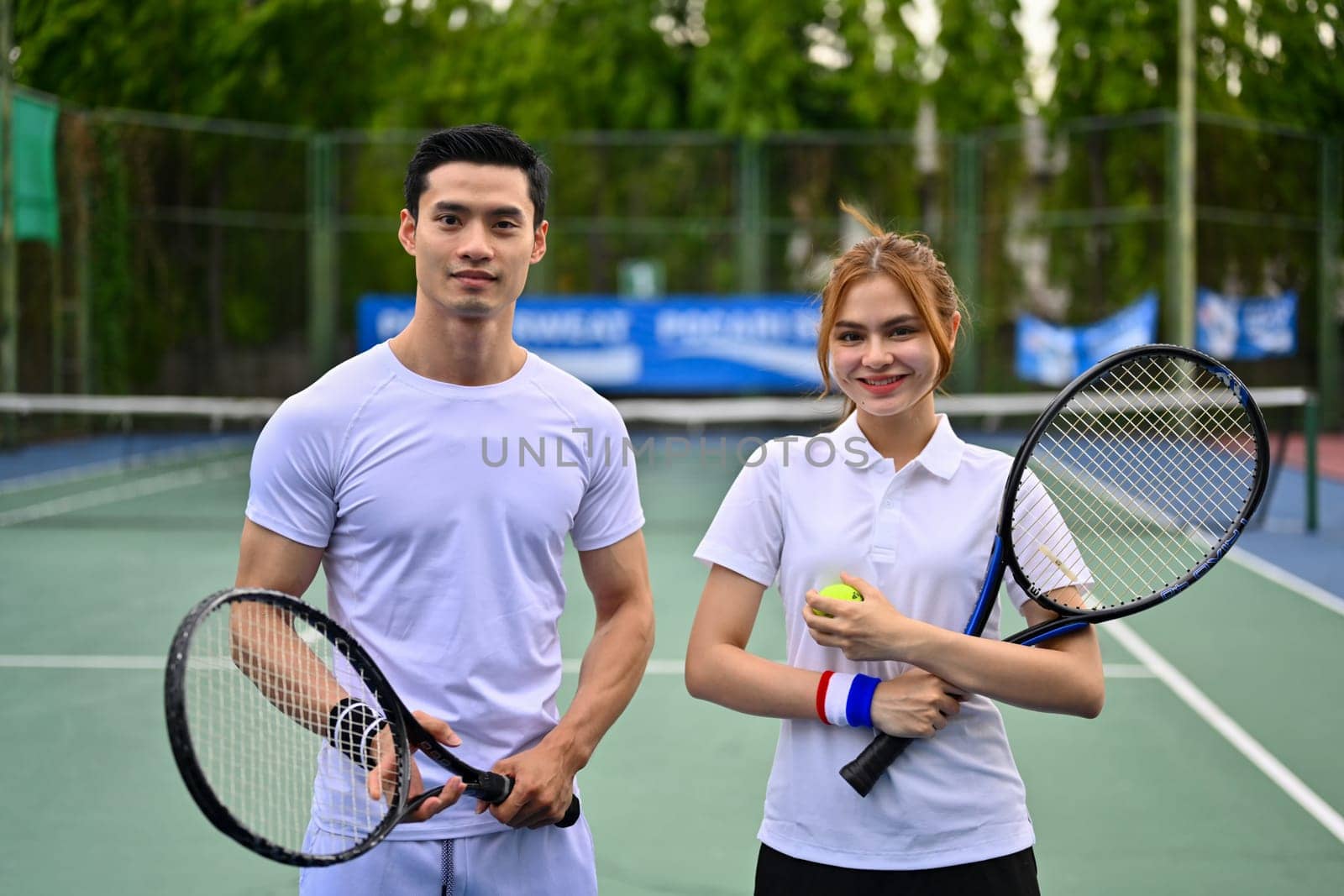 Two male and female tennis players standing at court on sunny day and smiling to camera by prathanchorruangsak