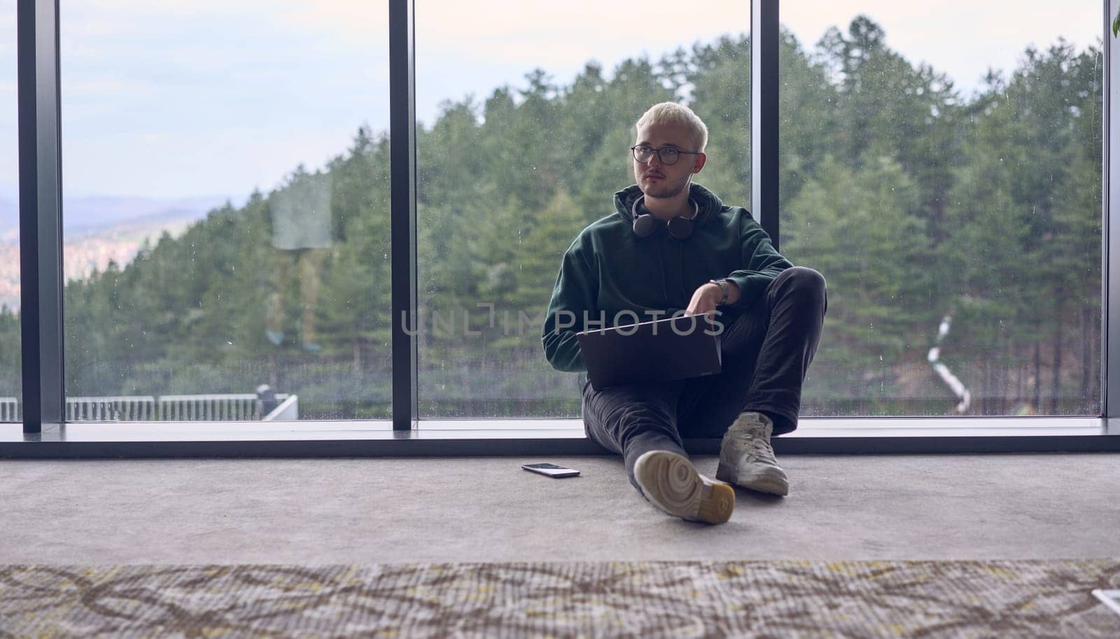 A young blond man in a modern office sits by the window, engrossed in his work on a laptop.
