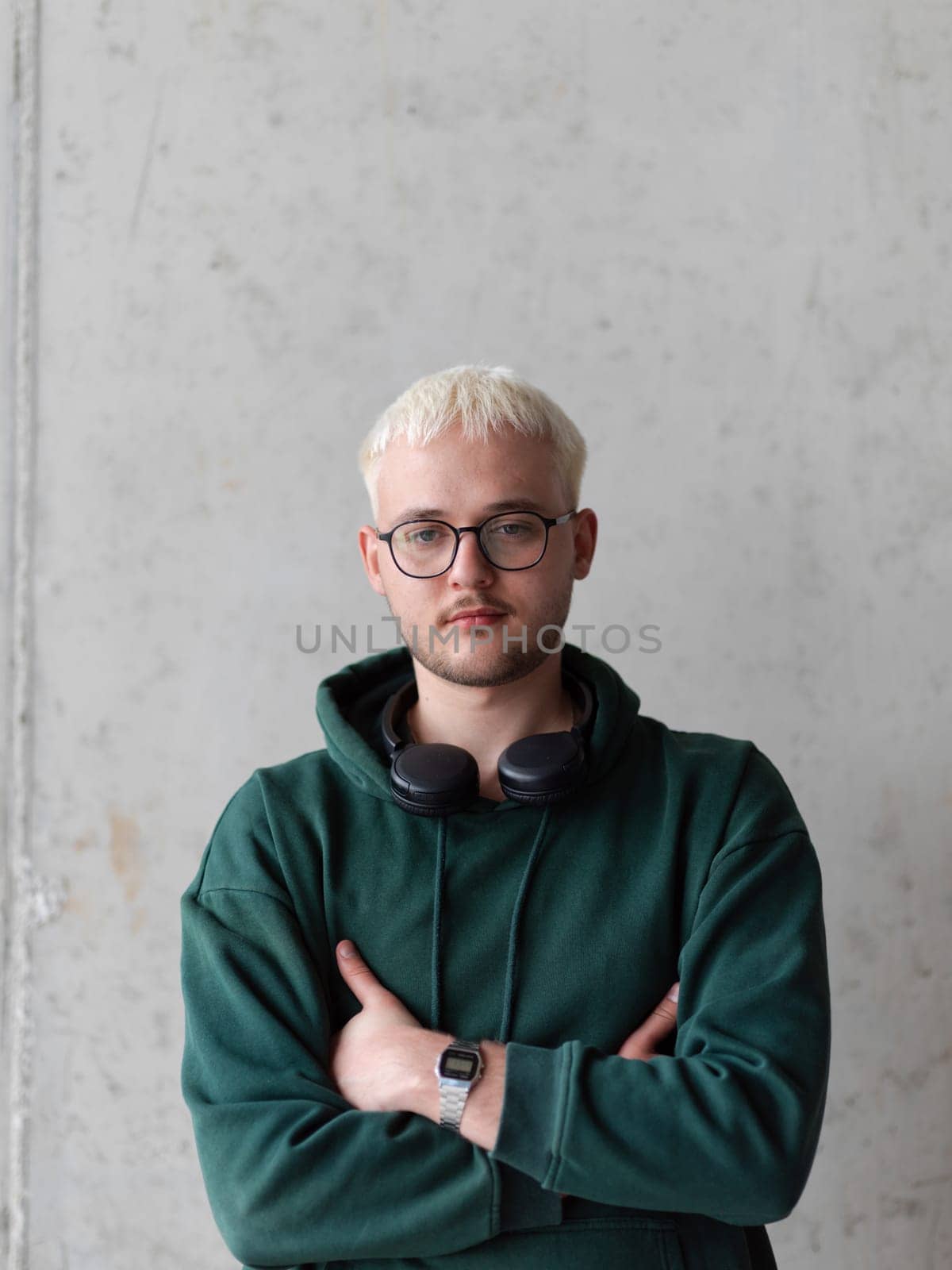 A man with blue hair, eyeglasses, and a green sweatshirt confidently poses with his arms crossed against a gray background, showcasing his fashionable and unique style