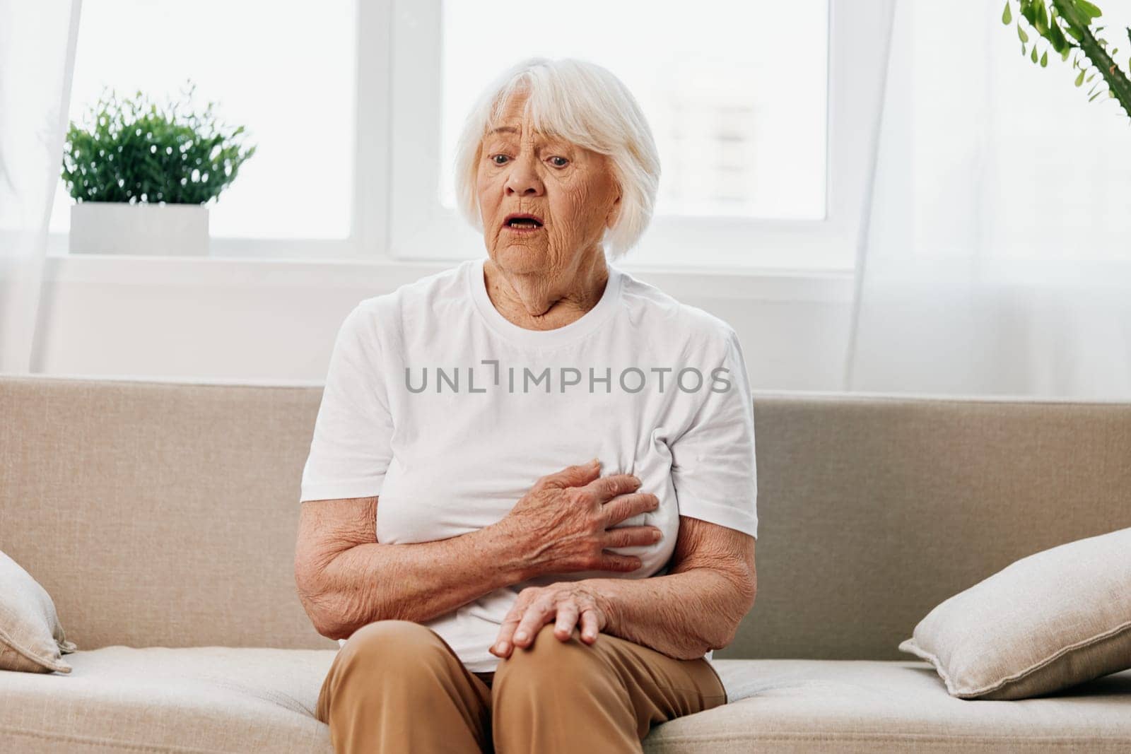 Elderly woman severe pain in the heart sitting on the couch, health problems in old age, poor quality of life. Grandmother with gray hair holding on to her chest, heart attack and stroke, pressure problem. High quality photo