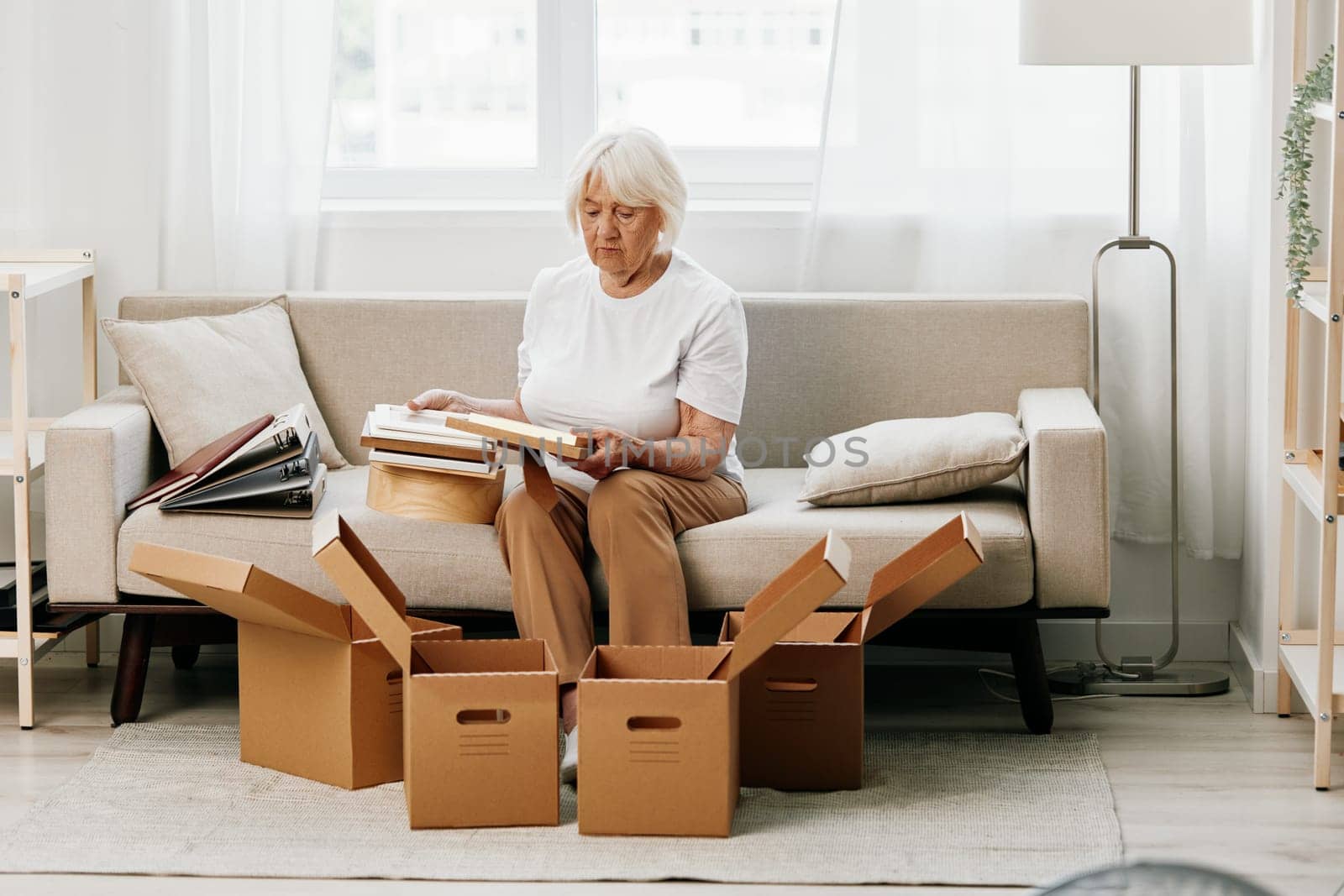 elderly woman sits on a sofa at home with boxes. collecting things with memories albums with photos and photo frames moving to a new place cleaning things and a happy smile. Lifestyle retirement. by SHOTPRIME