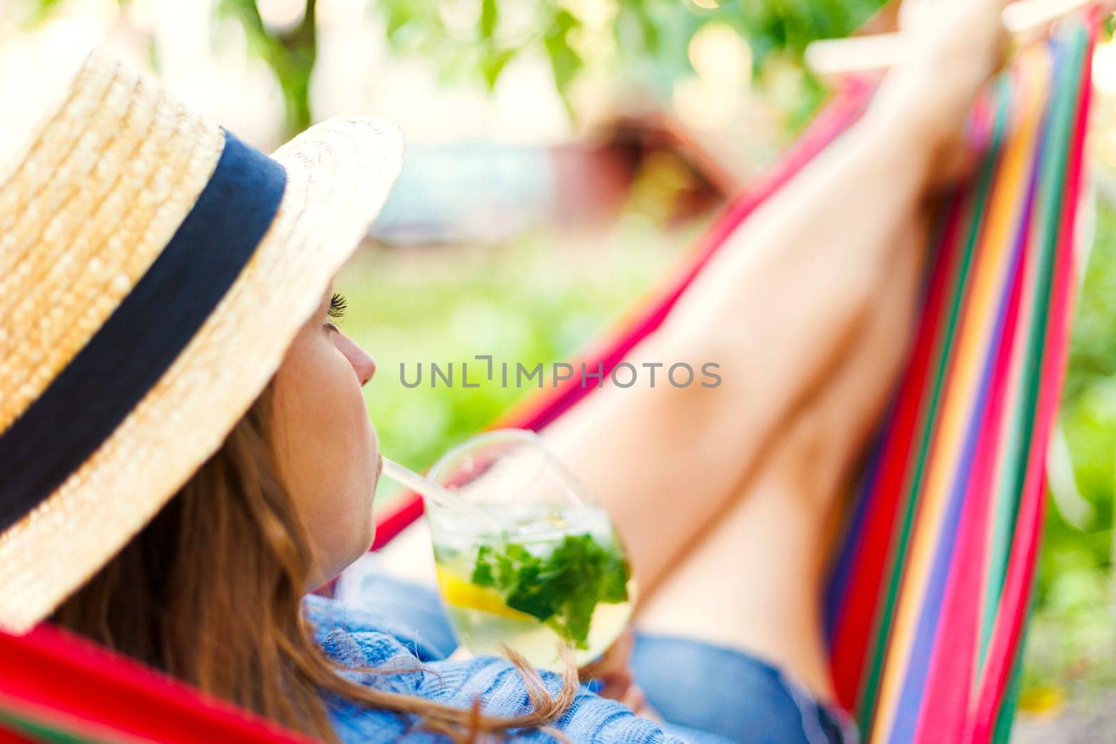 Young woman drinking a cocktail while lying in comfortable hammock at green garden.