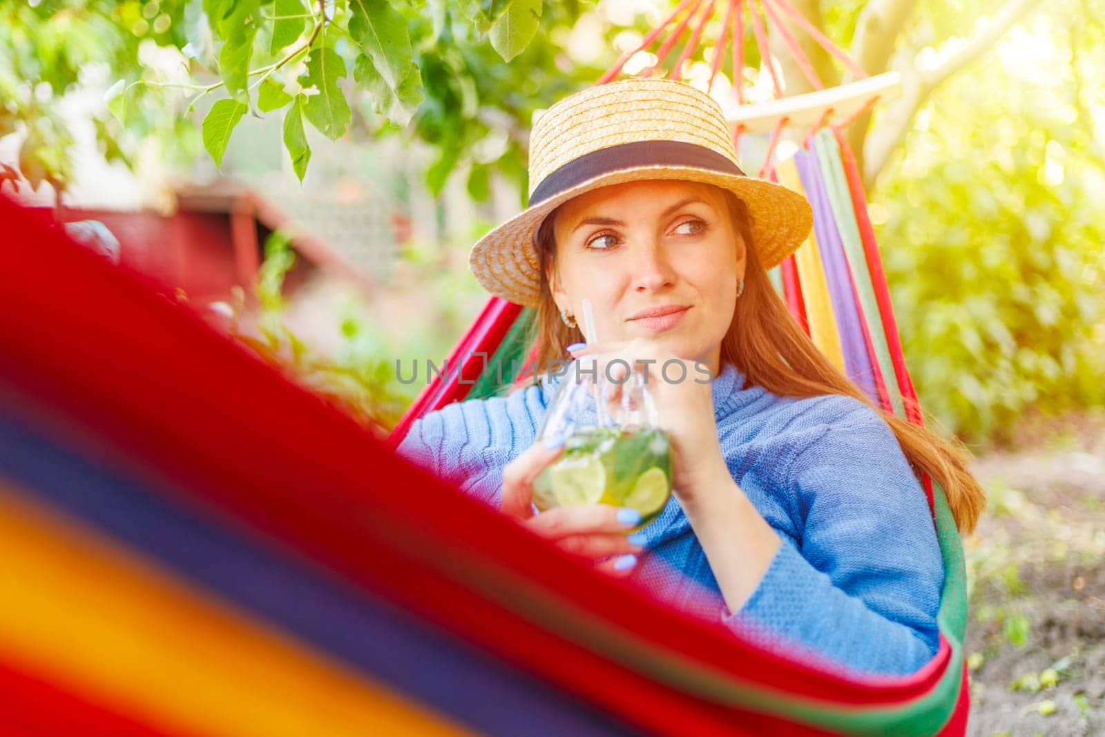 Young woman drinking a cocktail while lying in comfortable hammock at green garden.