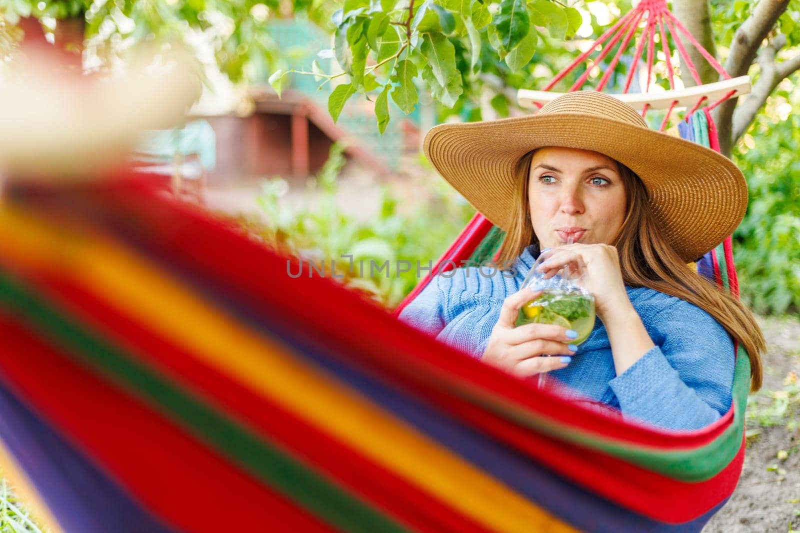 Young woman drinking a cocktail while lying in comfortable hammock at green garden.