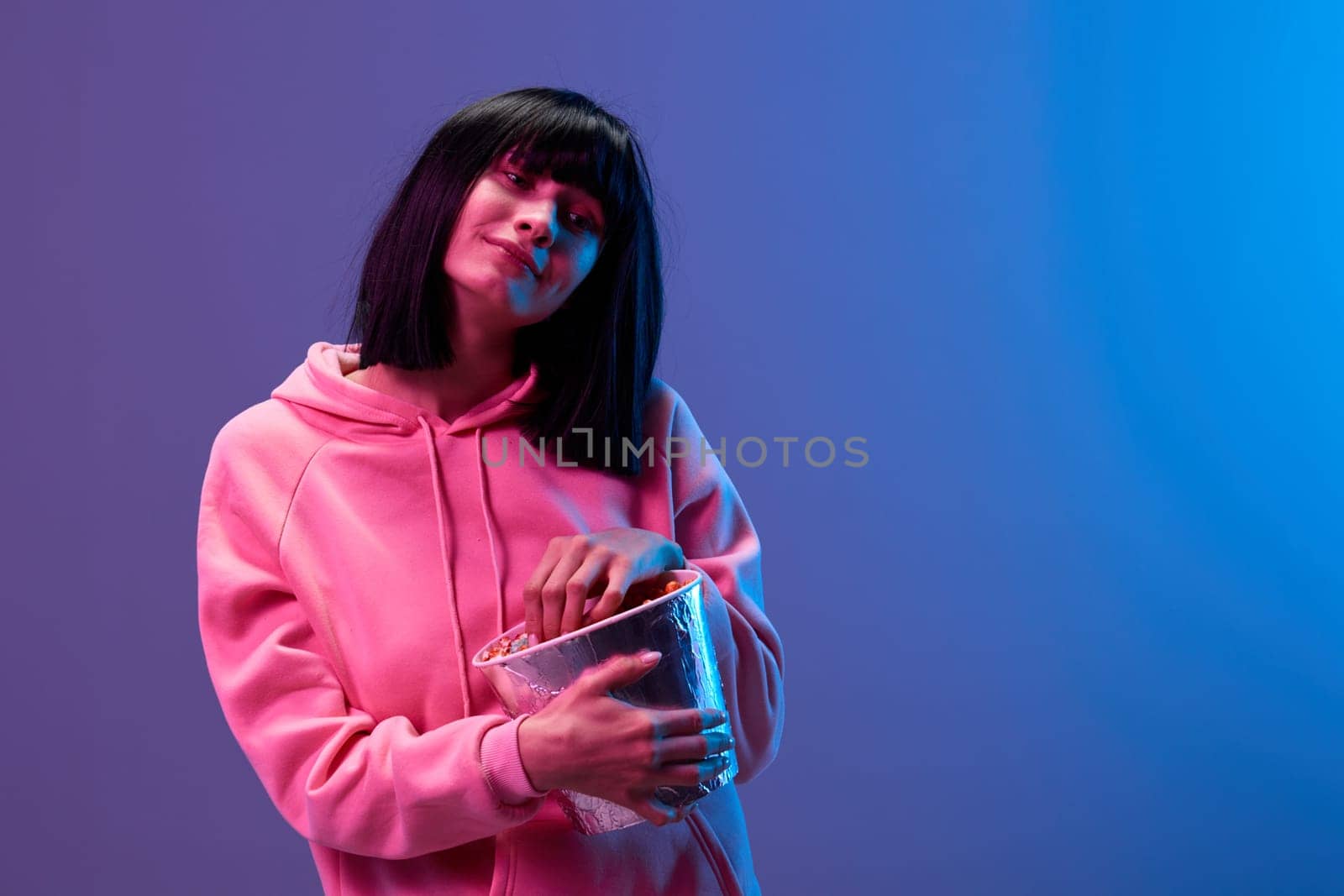Cute smiling awesome brunet woman in pink hoodie with popcorn look aside posing isolated in blue violet color light studio background. Neon party Cyberpunk Cinema concept. Copy space