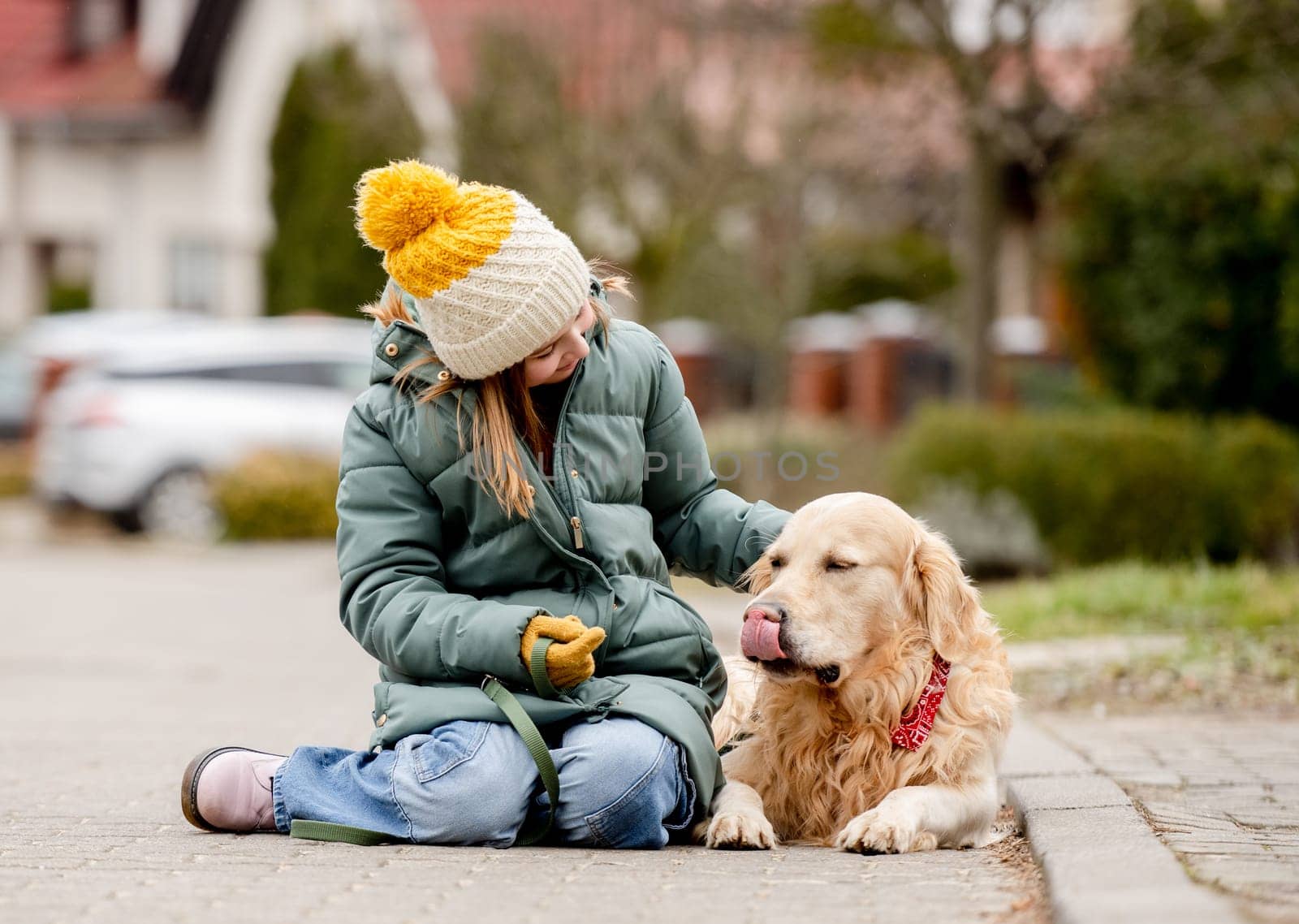 Preteen child girl sitting with golden retriever dog on asphalt at autumn street wearing hat and warm jacket. Pretty kid petting purebred pet doggy labrador outdoors at city