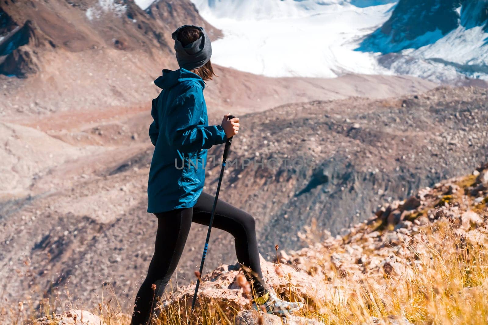 A young girl leads an active lifestyle, climbs a trail in the mountains with trekking poles, snow-capped mountain peaks and a glacier in the background.