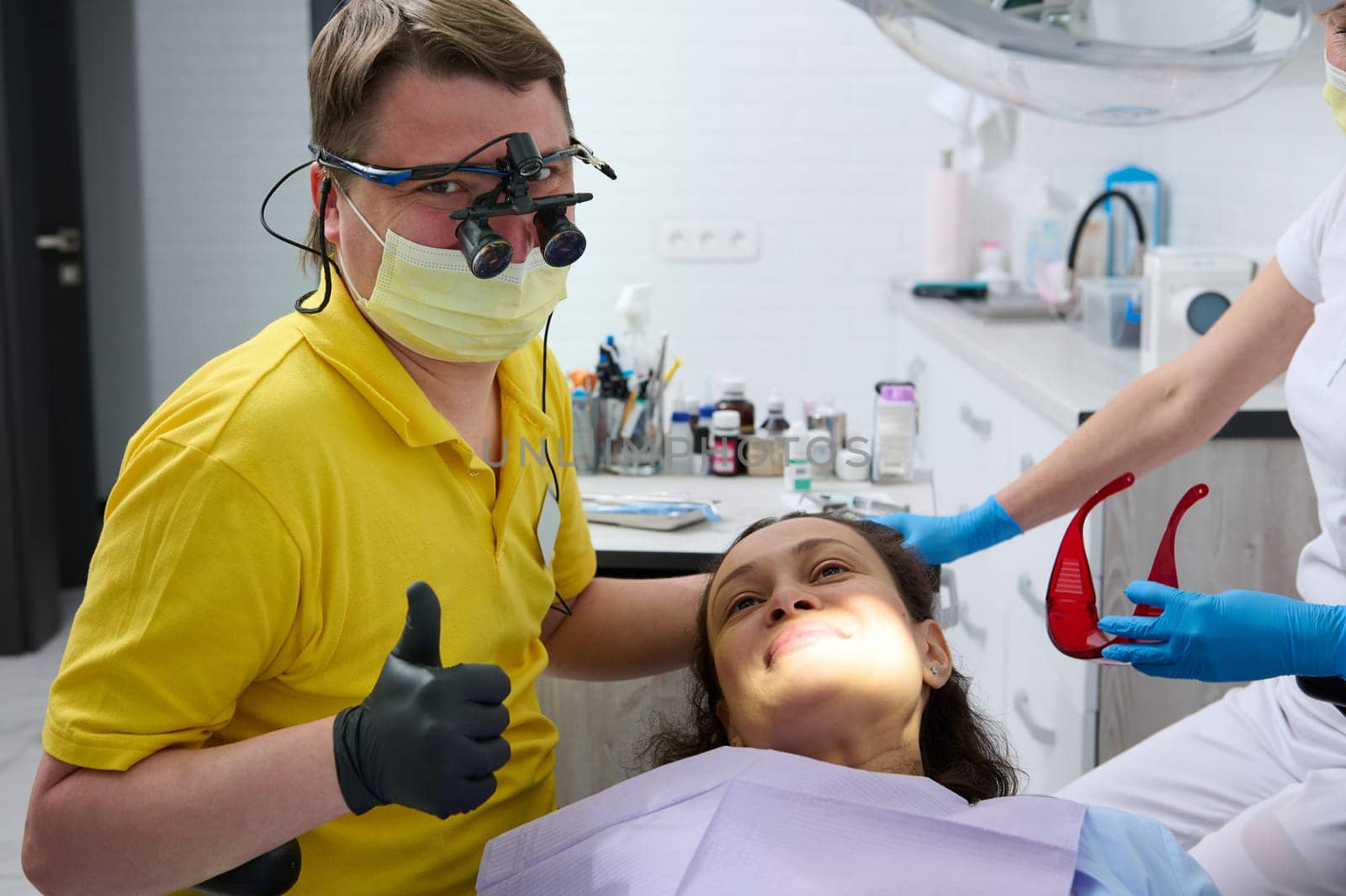 Happy professional dentist, hygienist orthodontist in protective medical mask and goggles, looking at camera while performing oral examination of female patient in dentistry clinic. Dental practice