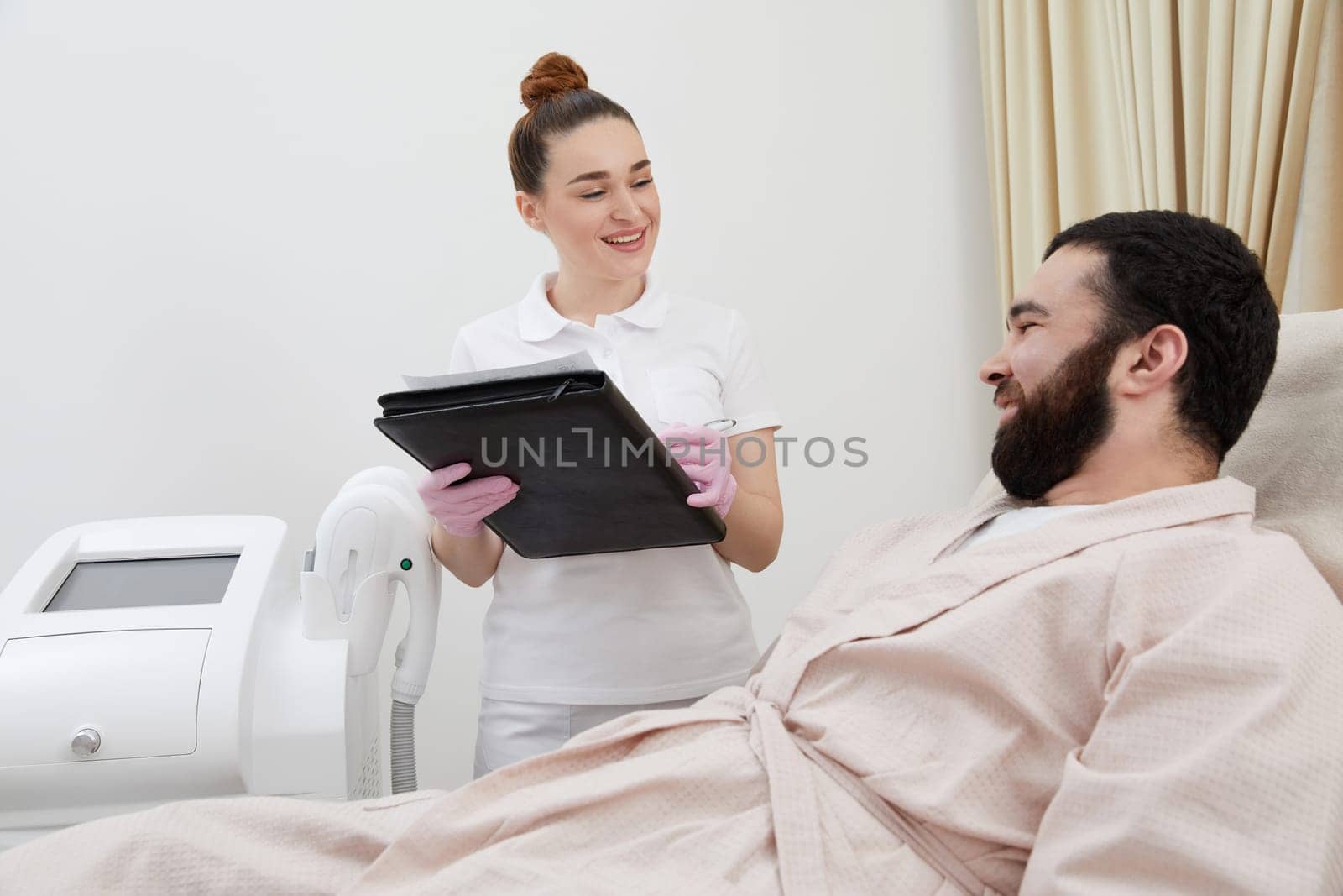 Professional cosmetician woman with papers talking to man before procedure in clinic of esthetic cosmetology by Mariakray