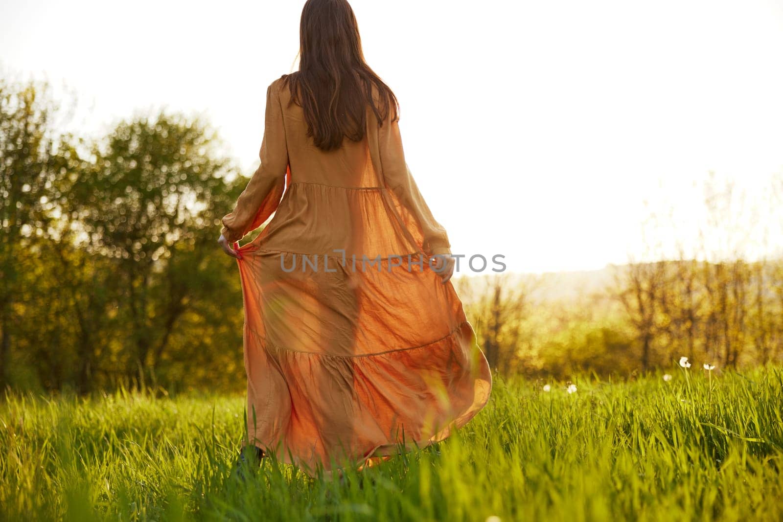 a slender woman stands in a long orange dress with her back to the camera illuminated by the sunset rays of the sun and looks towards the sky by Vichizh