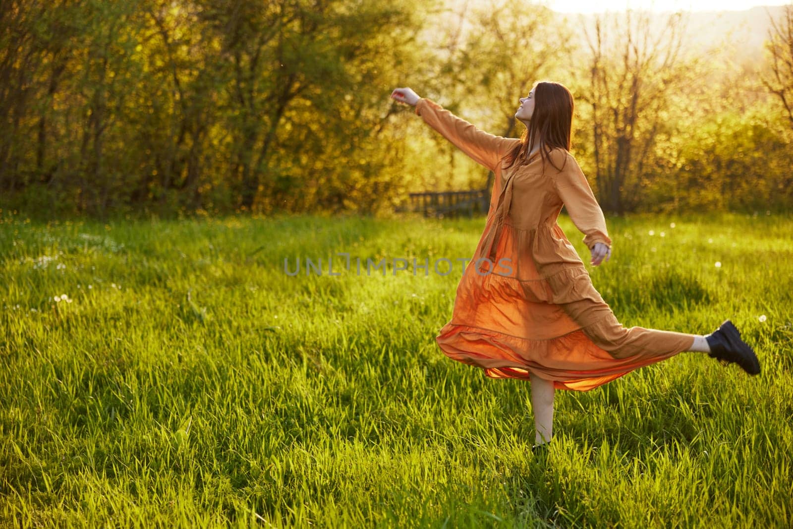 a happy woman in an orange dress illuminated by the rays of the setting sun happily jumps in a green field in the park, enjoying nature and a warm summer day. Horizontal photo. High quality photo