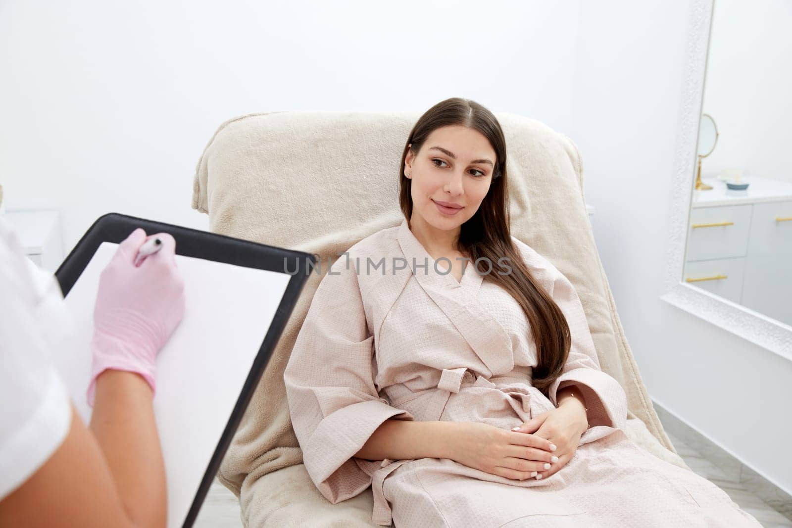 Smiling lady shows paper to woman client sitting in comfortable armchair in cosmetological clinic