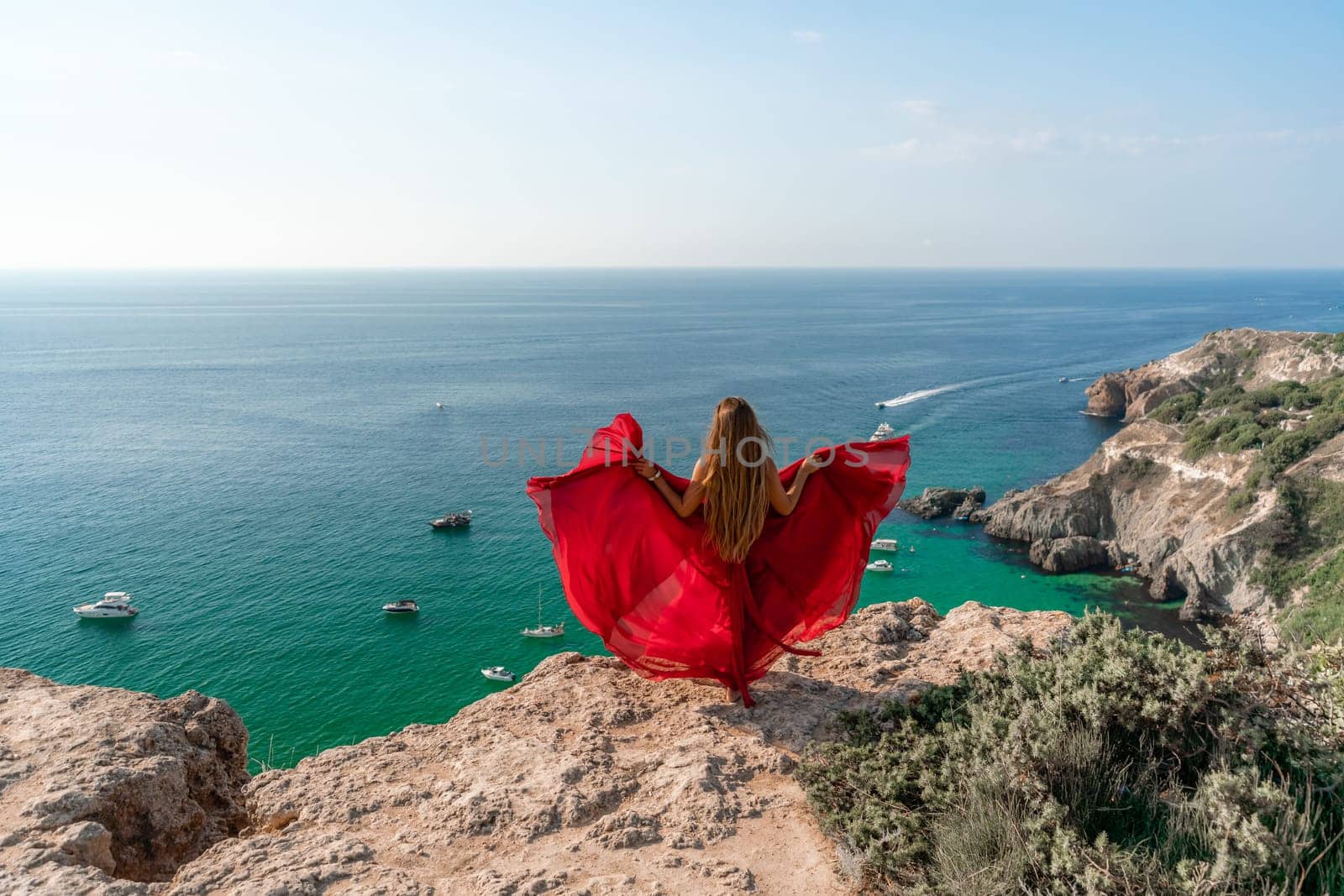 Woman sea red dress yachts. A beautiful woman in a red dress poses on a cliff overlooking the sea on a sunny day. Boats and yachts dot the background