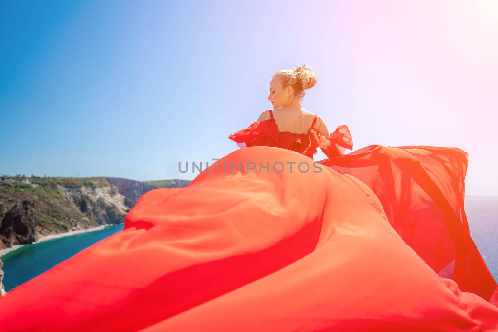 woman sea red dress. Blonde with long hair on a sunny seashore in a red flowing dress, back view, silk fabric waving in the wind. Against the backdrop of the blue sky and mountains on the seashore