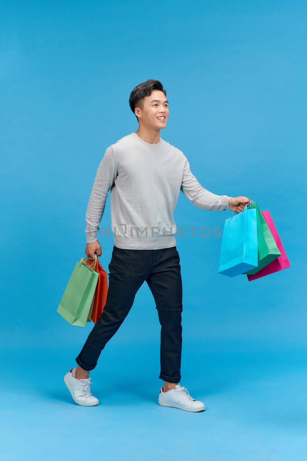 Young asian man shopaholic smiling while holding shopping bags isolated on blue by makidotvn
