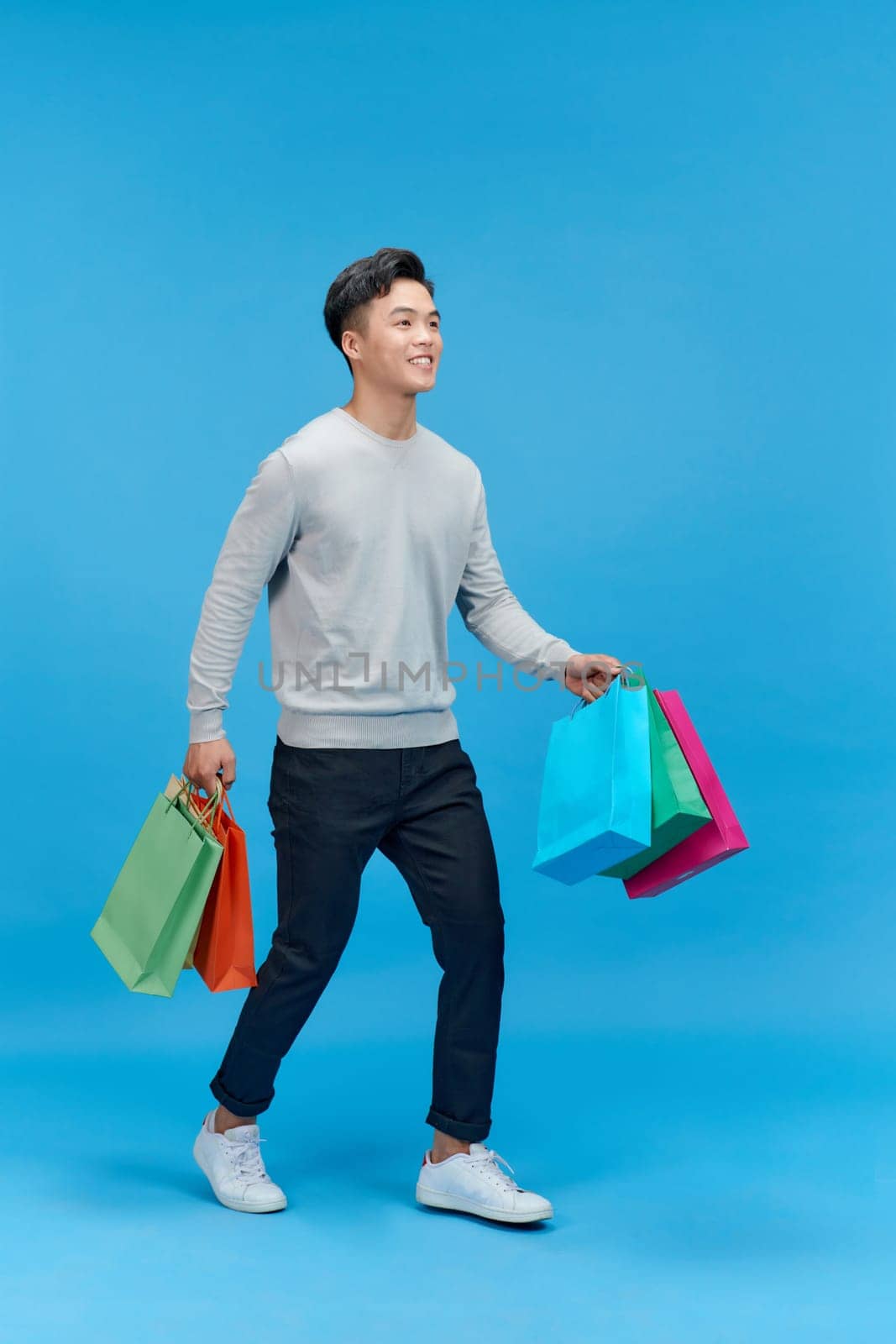 Young asian man shopaholic smiling while holding shopping bags isolated on blue