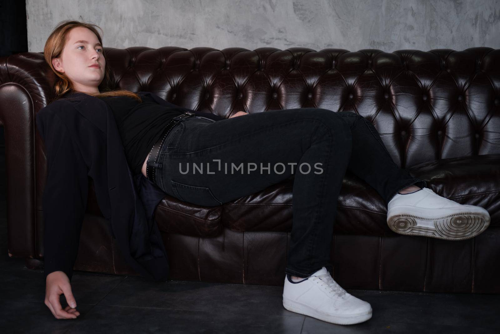 beautiful confident brunette woman in black on a brown leather couch in grey dark room. business woman. feminine. millennial people.