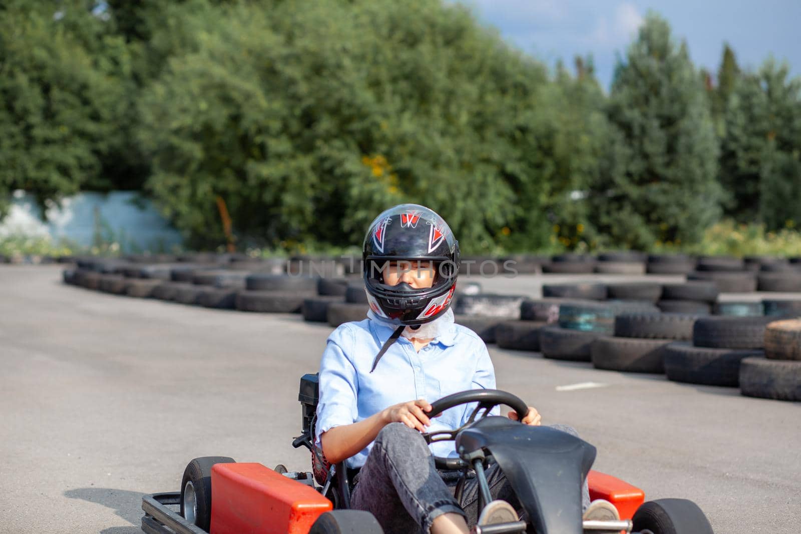 A girl or a woman in a hard hat rides a go-kart on a special track fenced with rubber wheels. Active recreation and sports on transport. Preparation and training for competitions. 