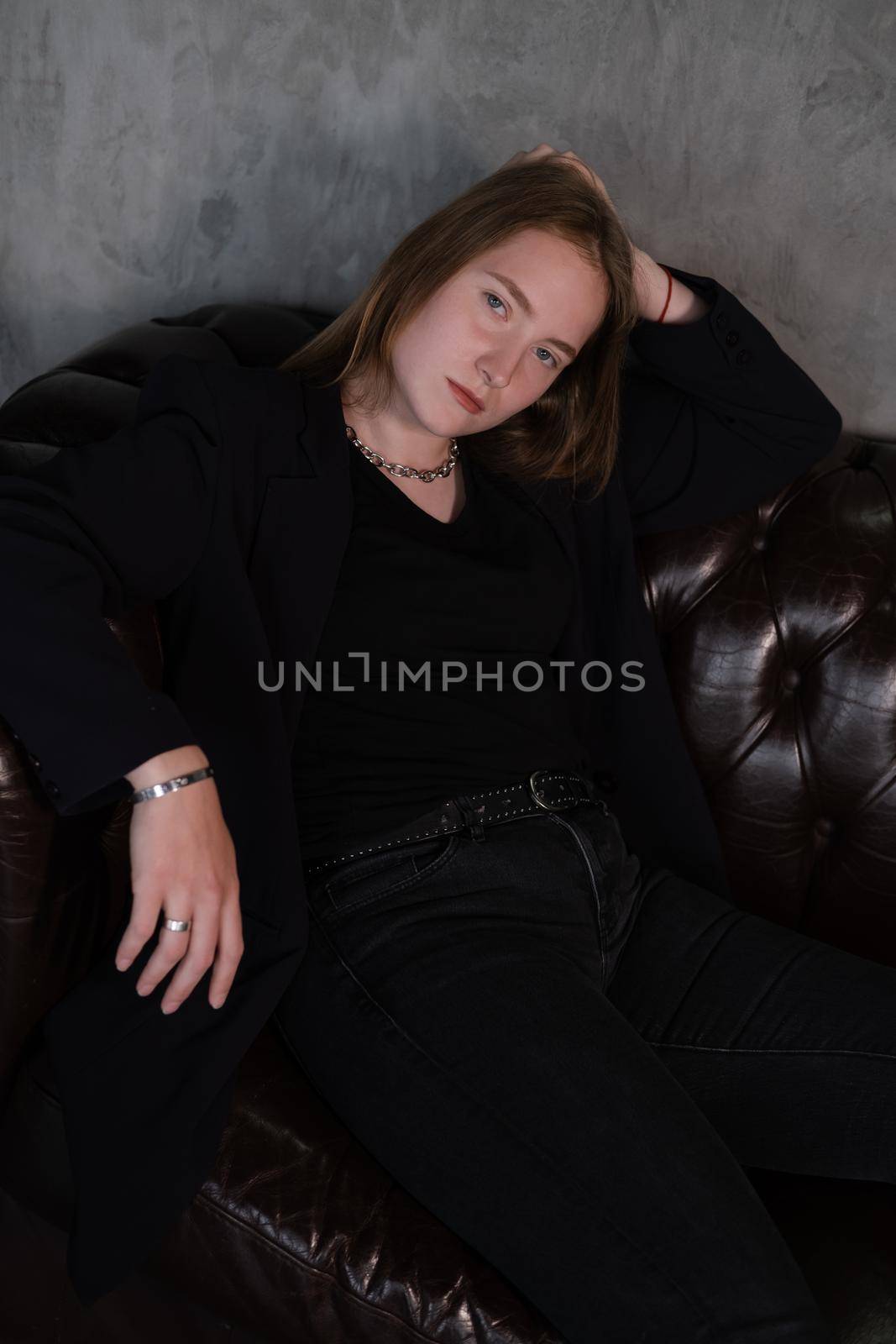 beautiful confident brunette woman in black on a brown leather couch in grey dark room. business woman. feminine. millennial people.