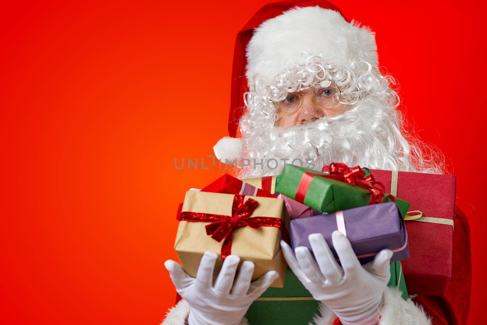 Happy Santa Claus with gift boxes in hands on red background with copy space. Christmas gifts concept