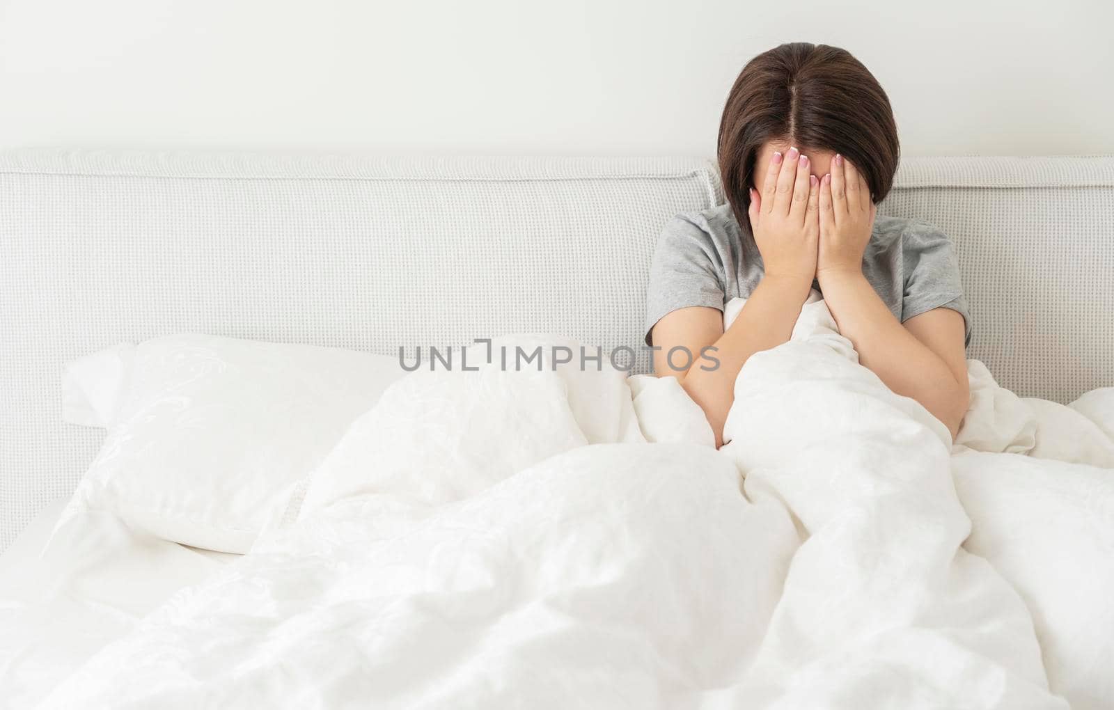 upset young brunette woman sitting on the white linen in bed at home, copyspace for your text to the left
