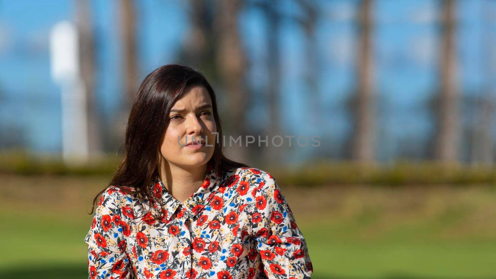 Portrait of beautiful young Latin woman looking to the right with floral design dress sitting in the middle of a garden with a background of trees during the morning