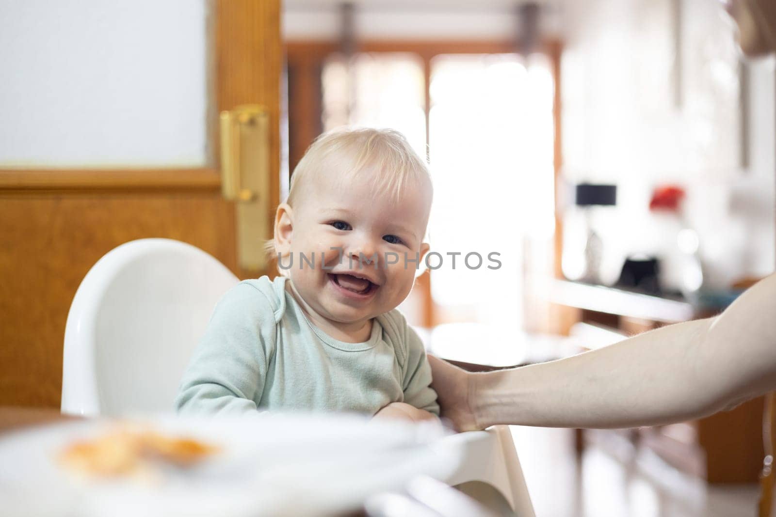 Adorable cheerful happy infant baby boy child smiling while sitting in high chair at the dining table in kitchen at home beeing spoon fed by his mother.