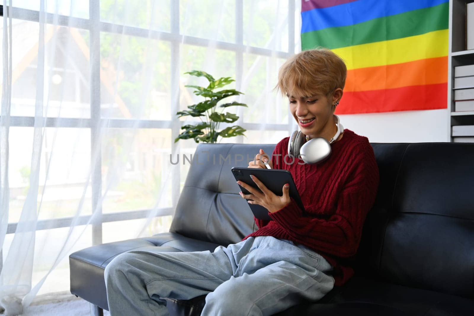 Positive teenage gay man in red sweater using digital tablet in bright living room with rainbow pride flag on background.