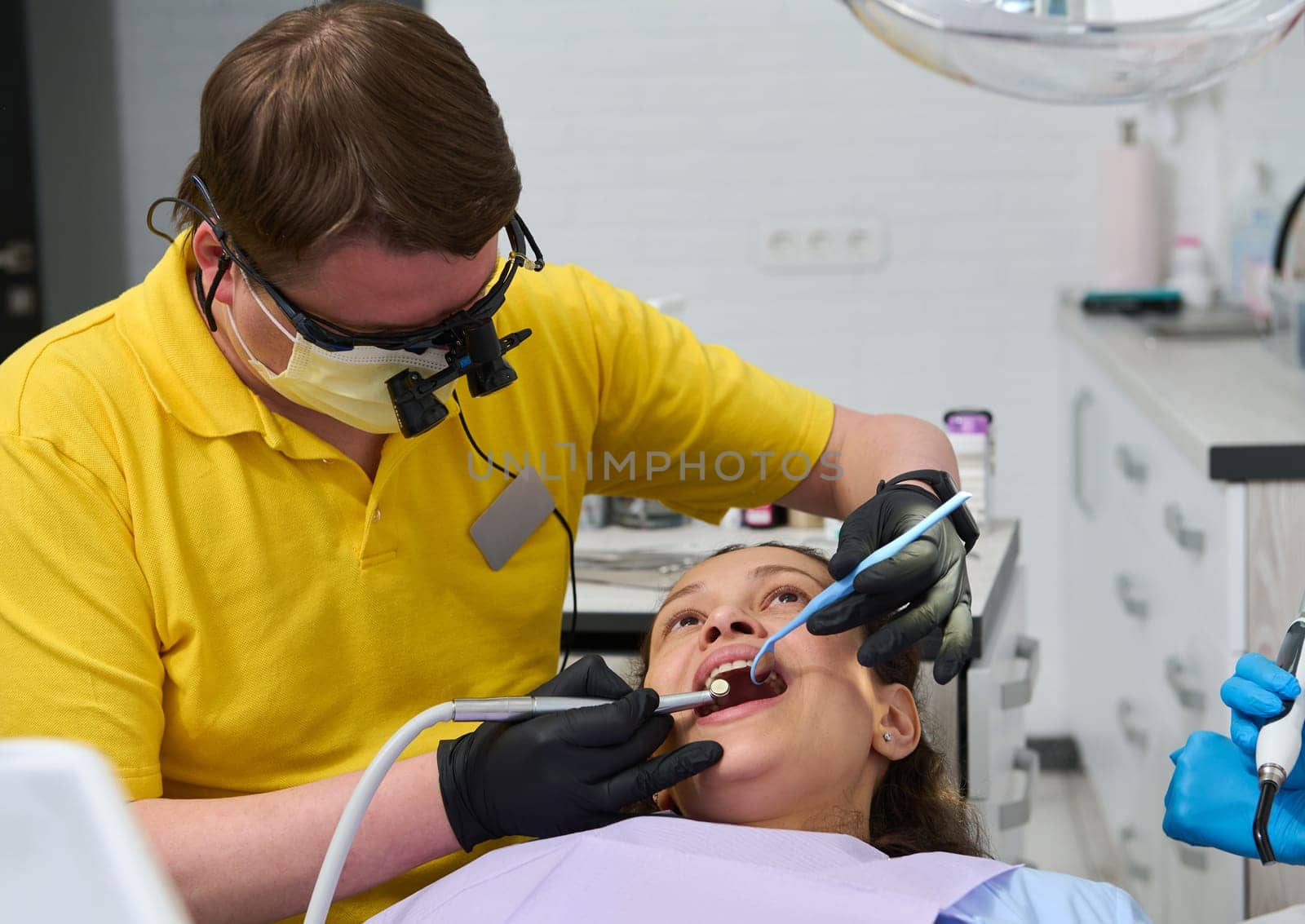 Professional dentist wearing orthodontist binoculars examines teeth and performs treatment to a patient in dental clinic by artgf