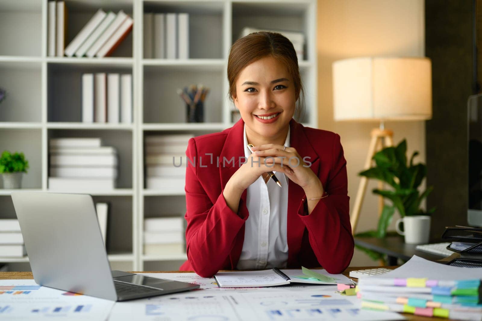 Elegant millennial executive woman in luxury suit sitting at modern workplace and smiling to camera.