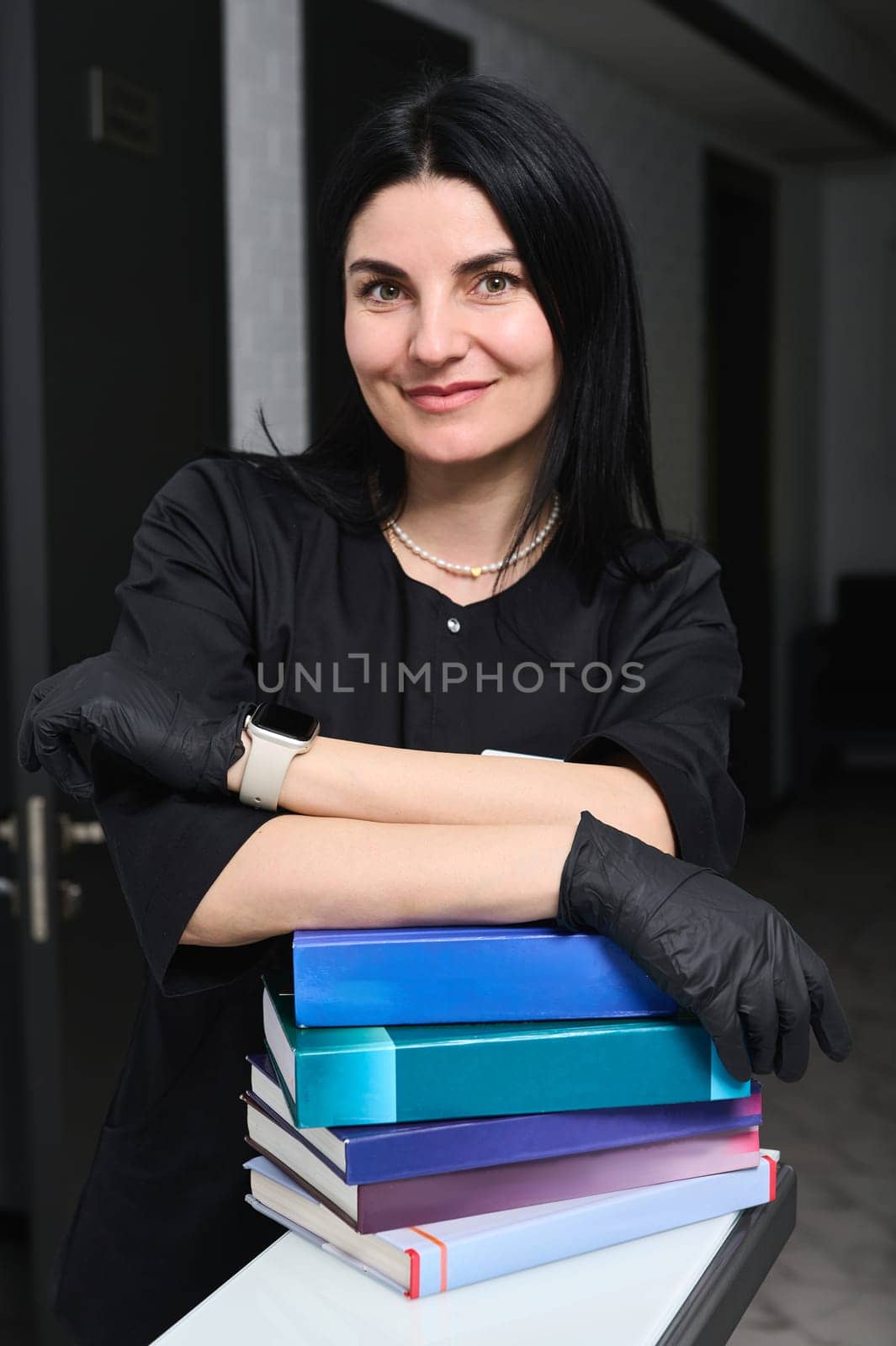 Authentic portrait of dark-haired Caucasian attractive confident experienced woman doctor, dressed in black medical uniform, standing at desk with educational books, smiles looking at camera. Medicine