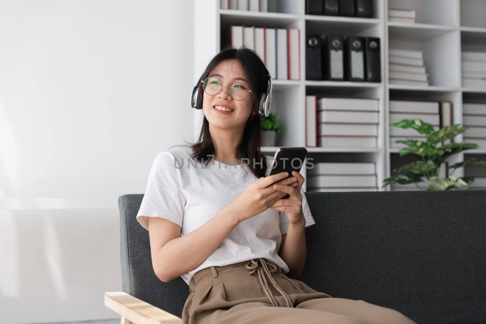 Young Asians woman listening to music on couch in living room at home. Happy Asia female using mobile smartphone, wearing headset and sitting on sofa.
