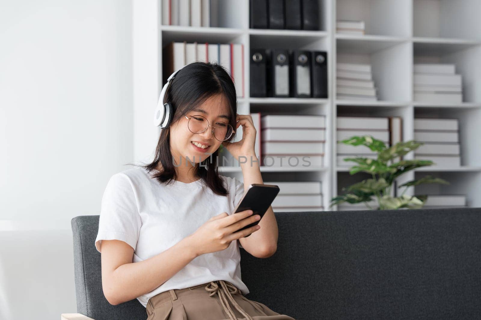 Young Asians woman listening to music on couch in living room at home. Happy Asia female using mobile smartphone, wearing headset and sitting on sofa.