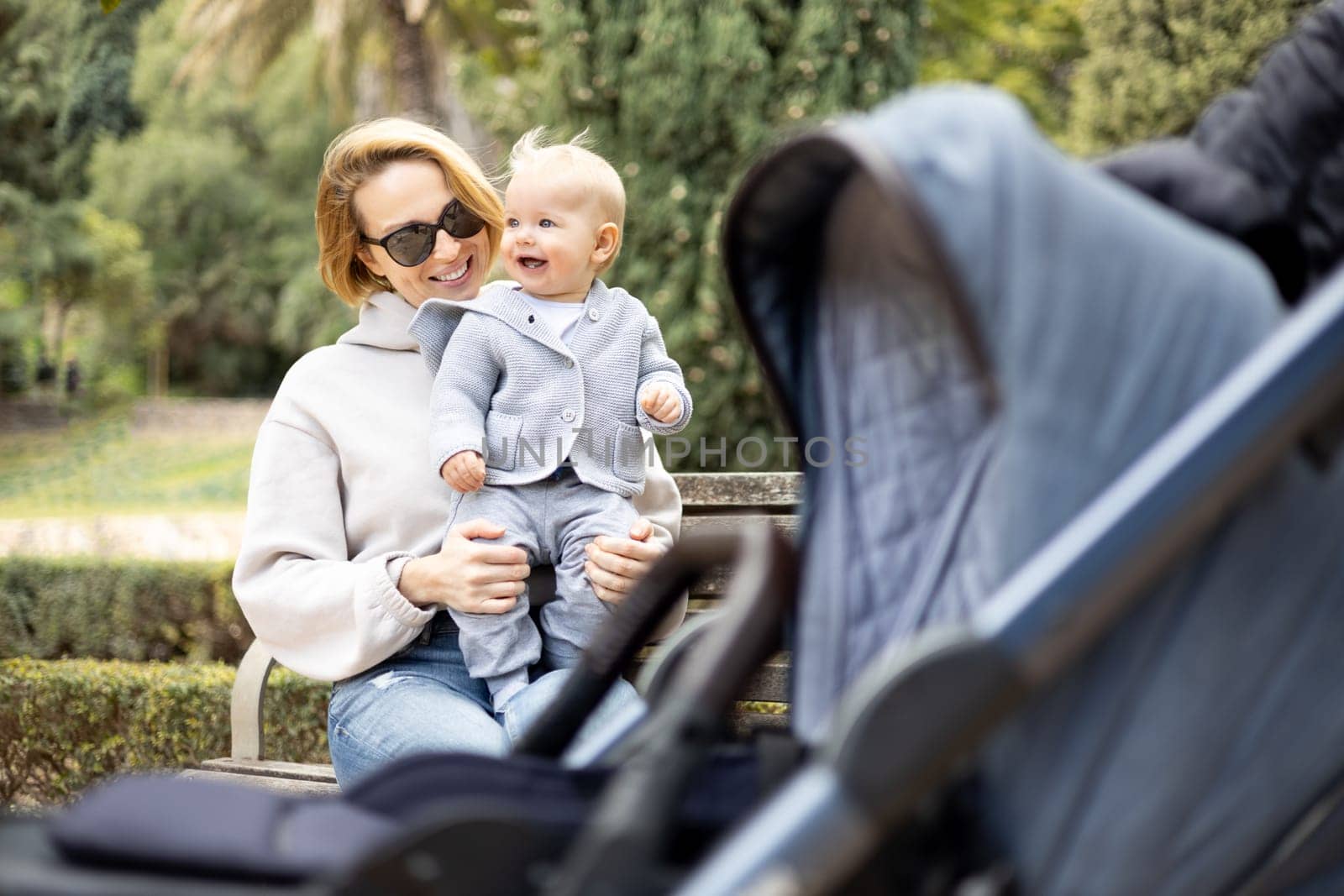Mother sitting on bench in urban park, laughing cheerfully, holding her smiling infant baby boy child in her lap having baby stroller parked by their site