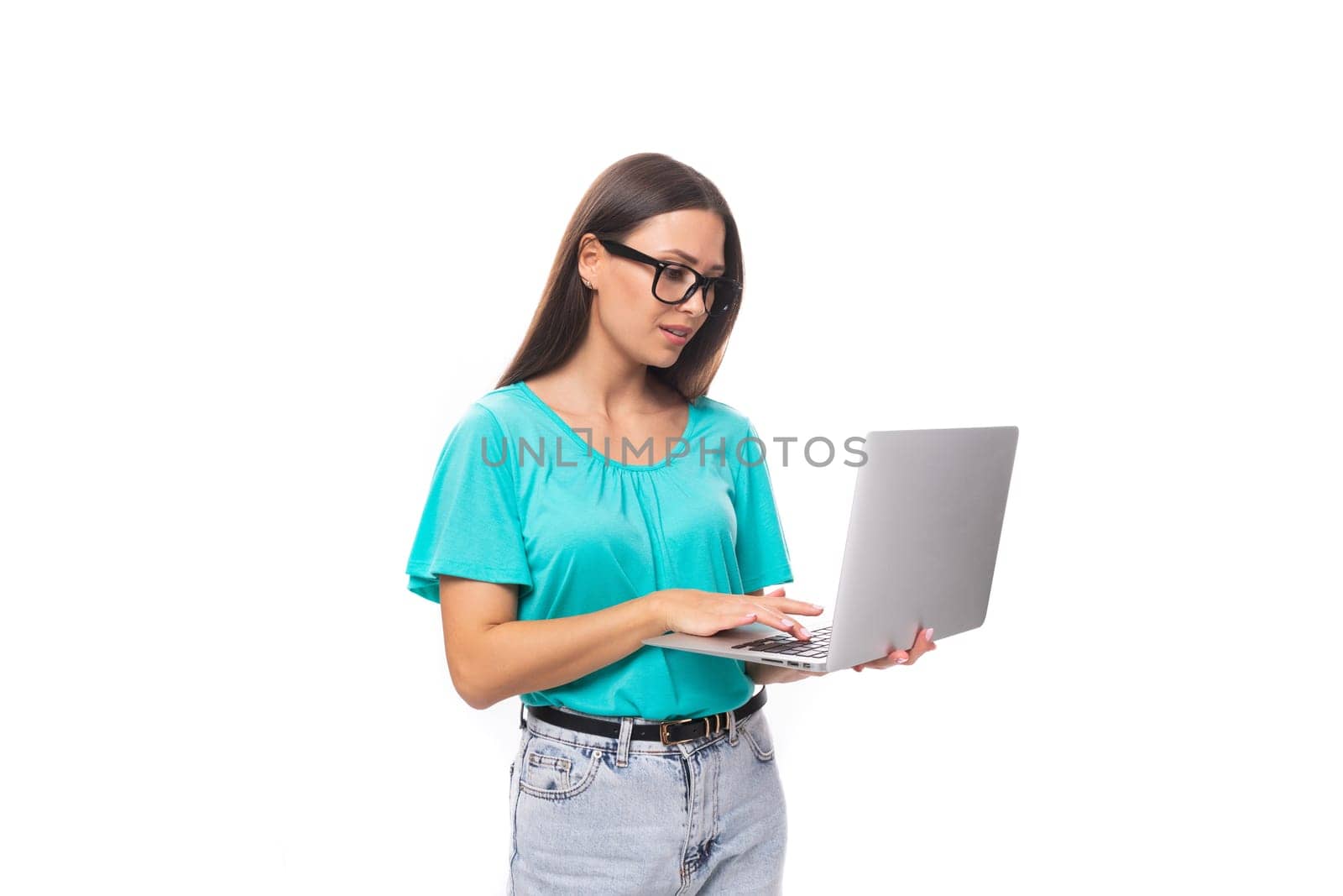 young smart caucasian female model in glasses with black silky smooth hair dressed in a blue t-shirt works using the internet and a laptop.