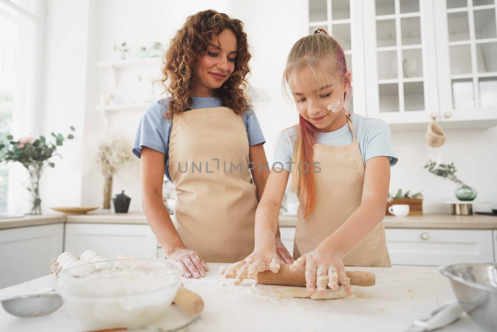 Teen girl helping her mom to cook dough in their kitchen at home by Fabrikasimf