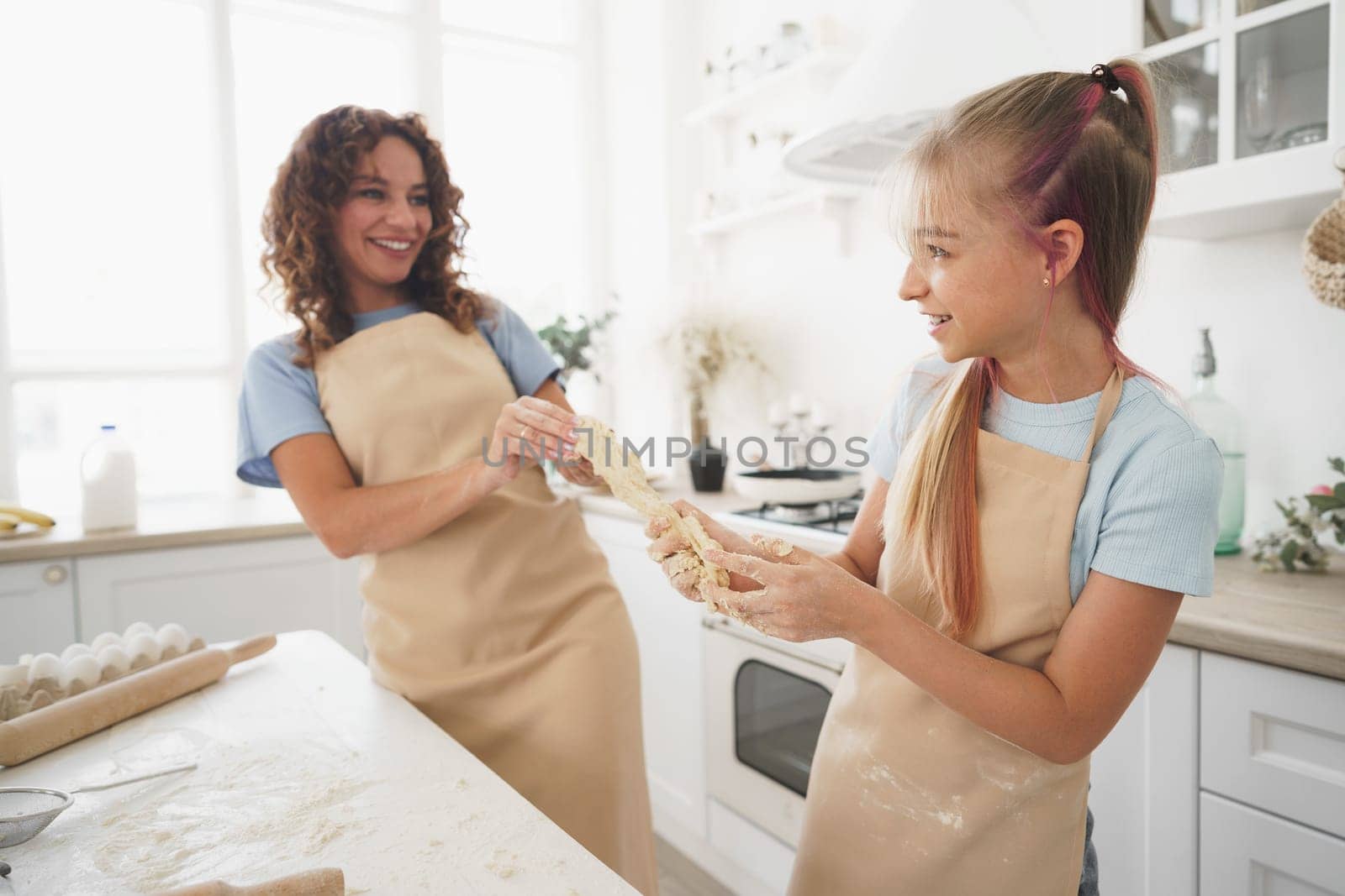 Teen girl helping her mom to cook dough in their kitchen at home by Fabrikasimf