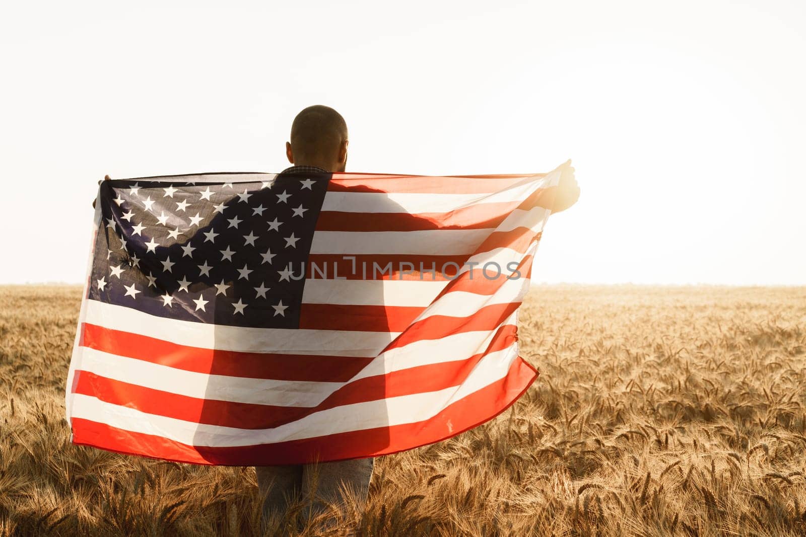 Young man holding American flag on back while standing in wheat field on dawn