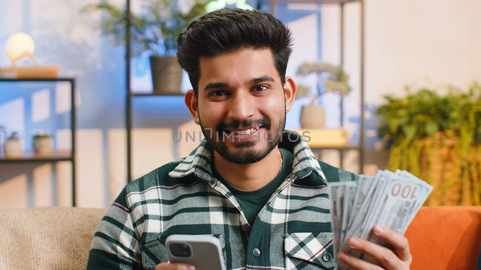 Planning family budget. Smiling indian man counting money cash, use smartphone calculate domestic bills at home room. Joyful hindu guy satisfied of income and saves money for planned vacation, gifts