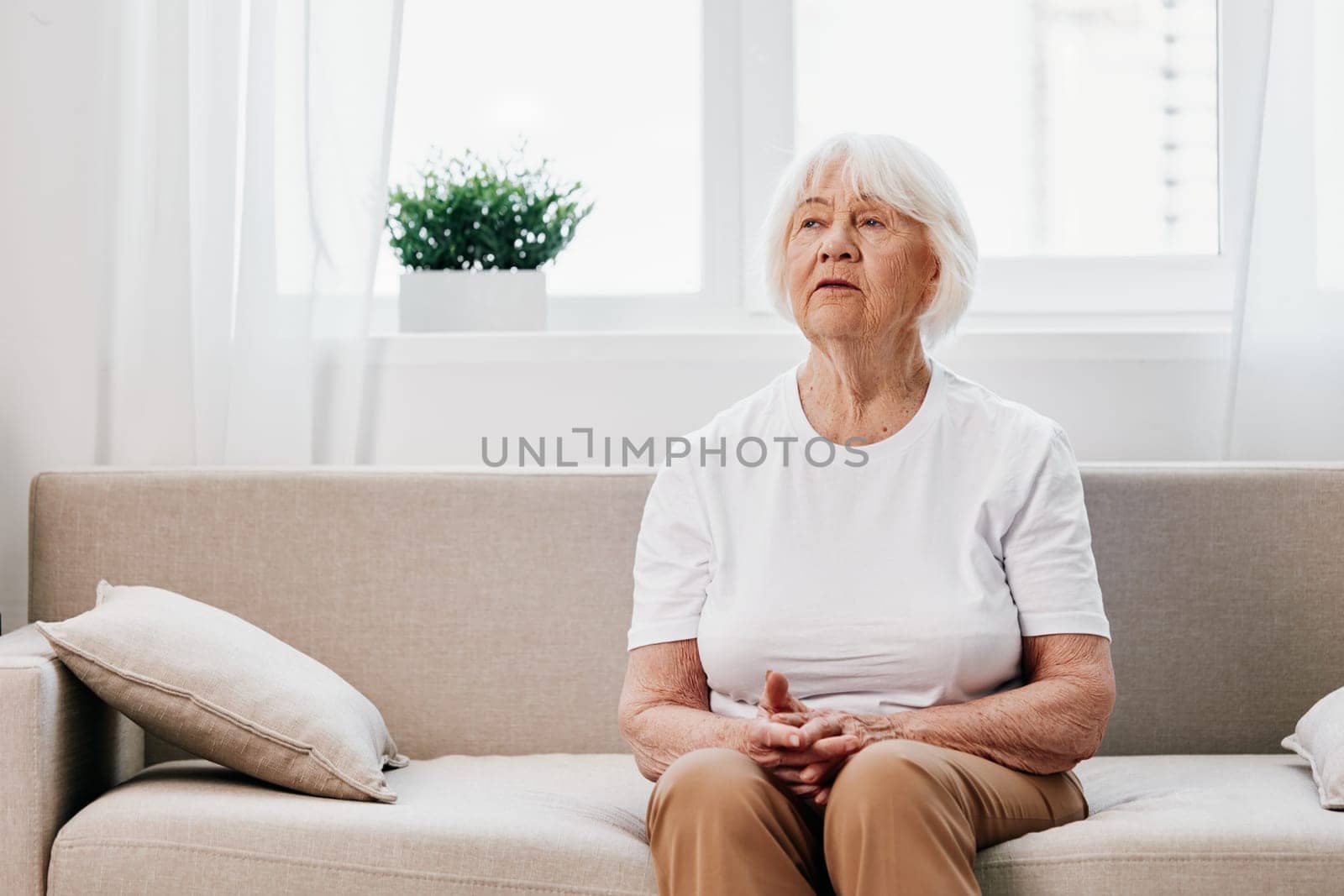 Elderly woman sits on sofa at home, bright spacious interior in old age smile, lifestyle. Grandmother with gray hair in a white T-shirt and beige trousers. High quality photo